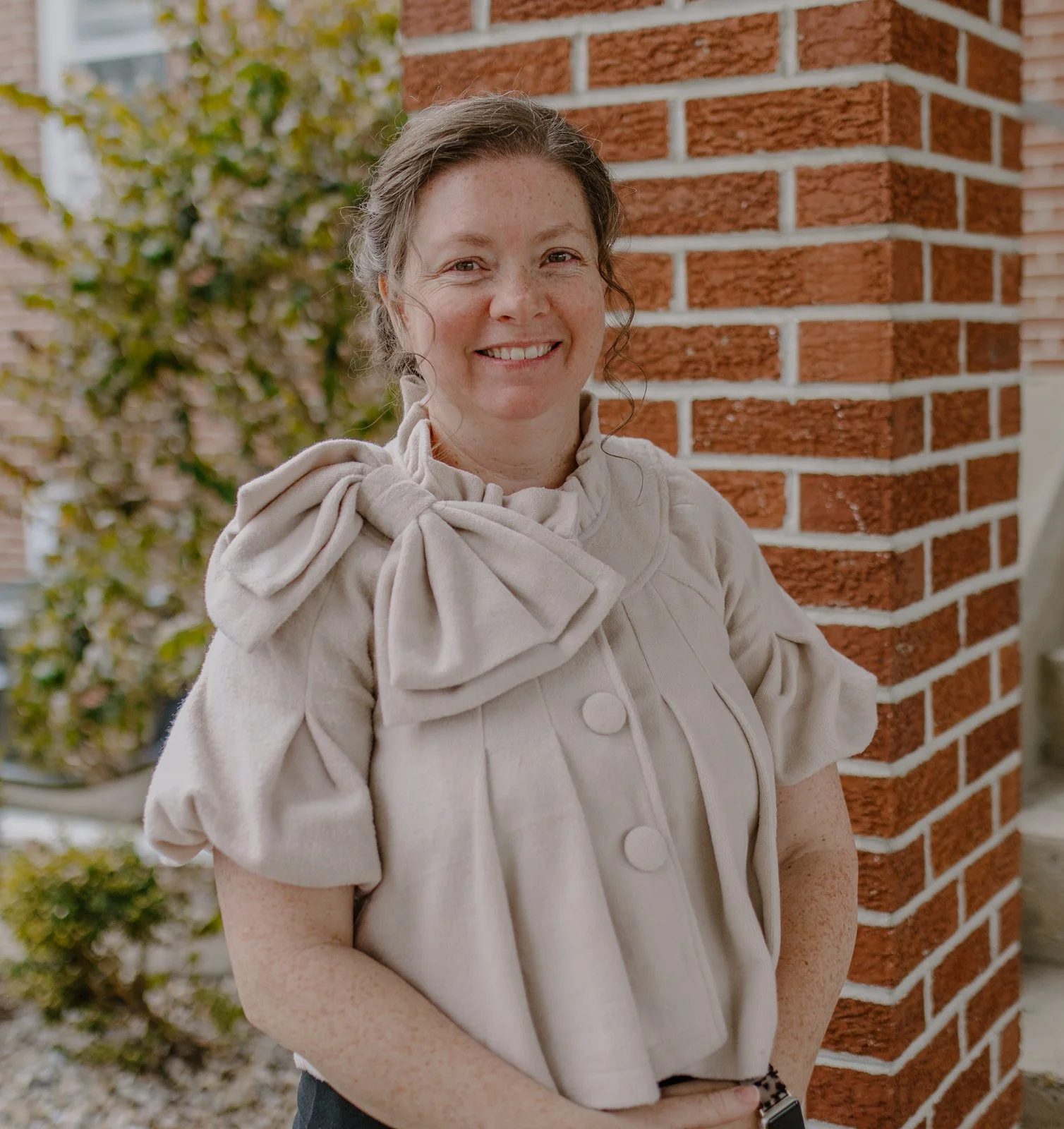A woman with curly brown hair and freckles, smiling, wearing a beige coat with large bow details on the shoulders, standing outdoors next to a brick pillar.