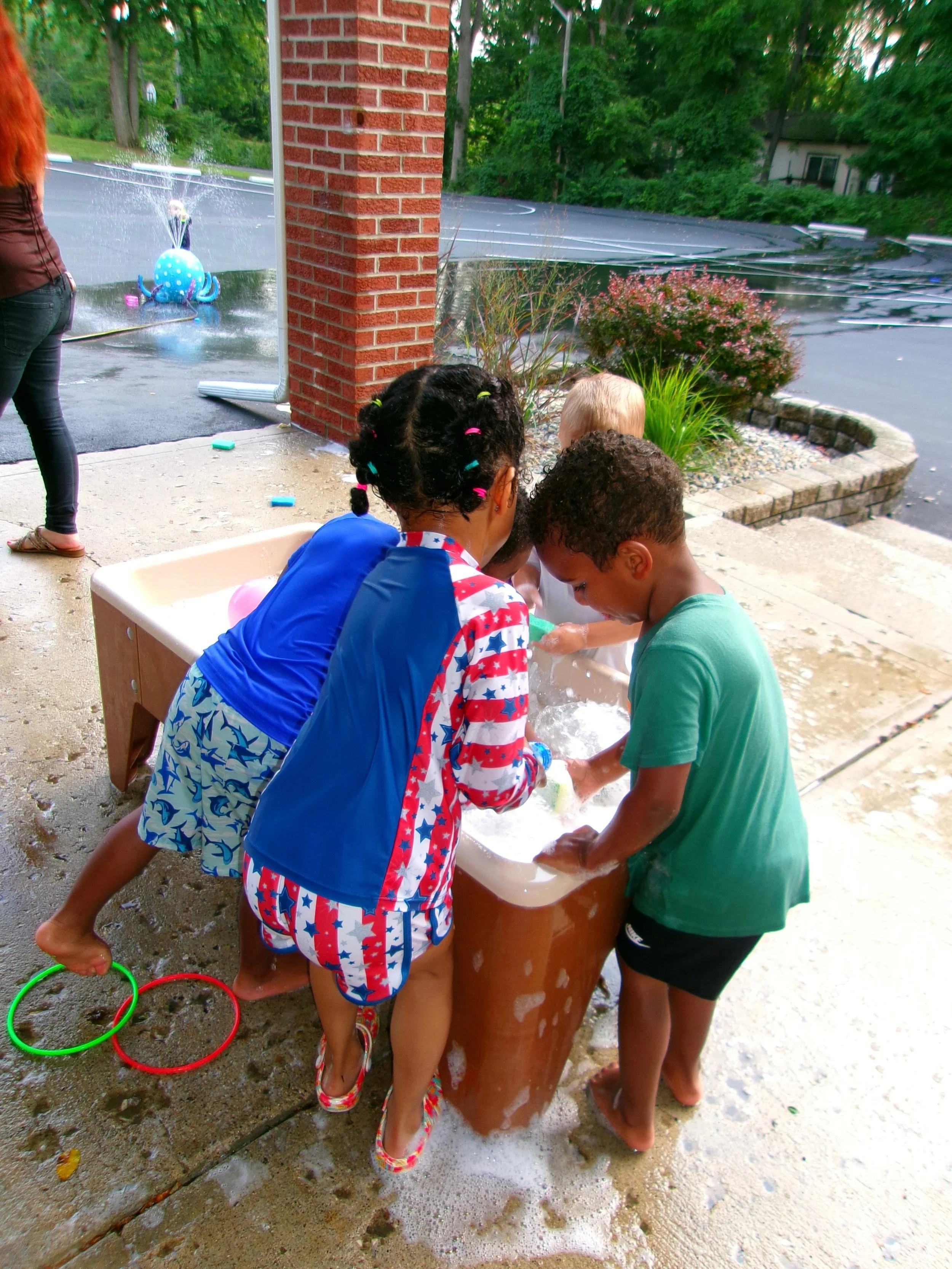 Children gathered around a water table, playing and washing their hands with soap outdoors. There is a brick wall and lush greenery in the background.