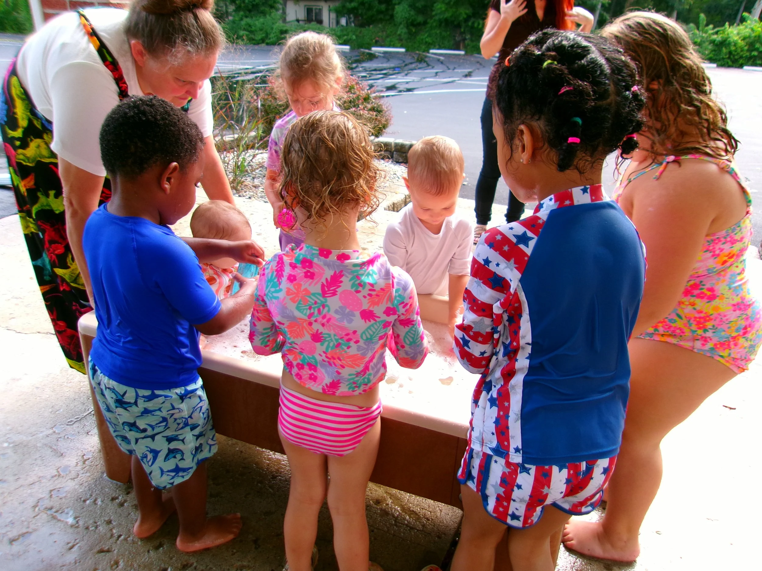Children and adults gathered around an outdoor table, looking at an object on the table, with greenery and parking lot in the background.
