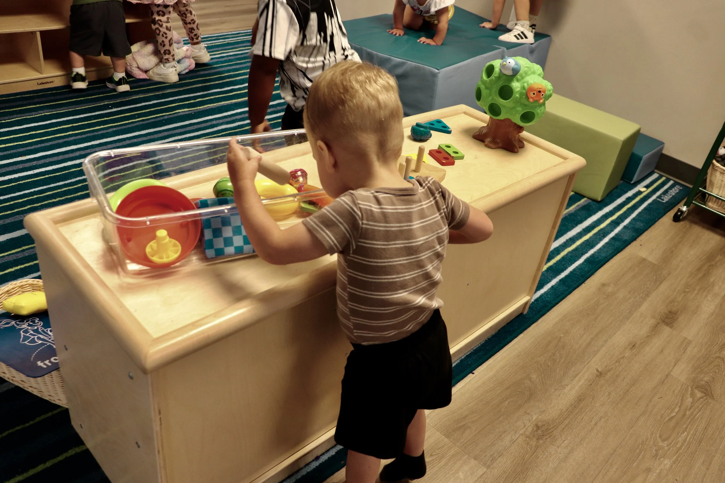 Children playing and exploring toys in a preschool or daycare setting, with colorful toys on a wooden table and children on mats in the background.
