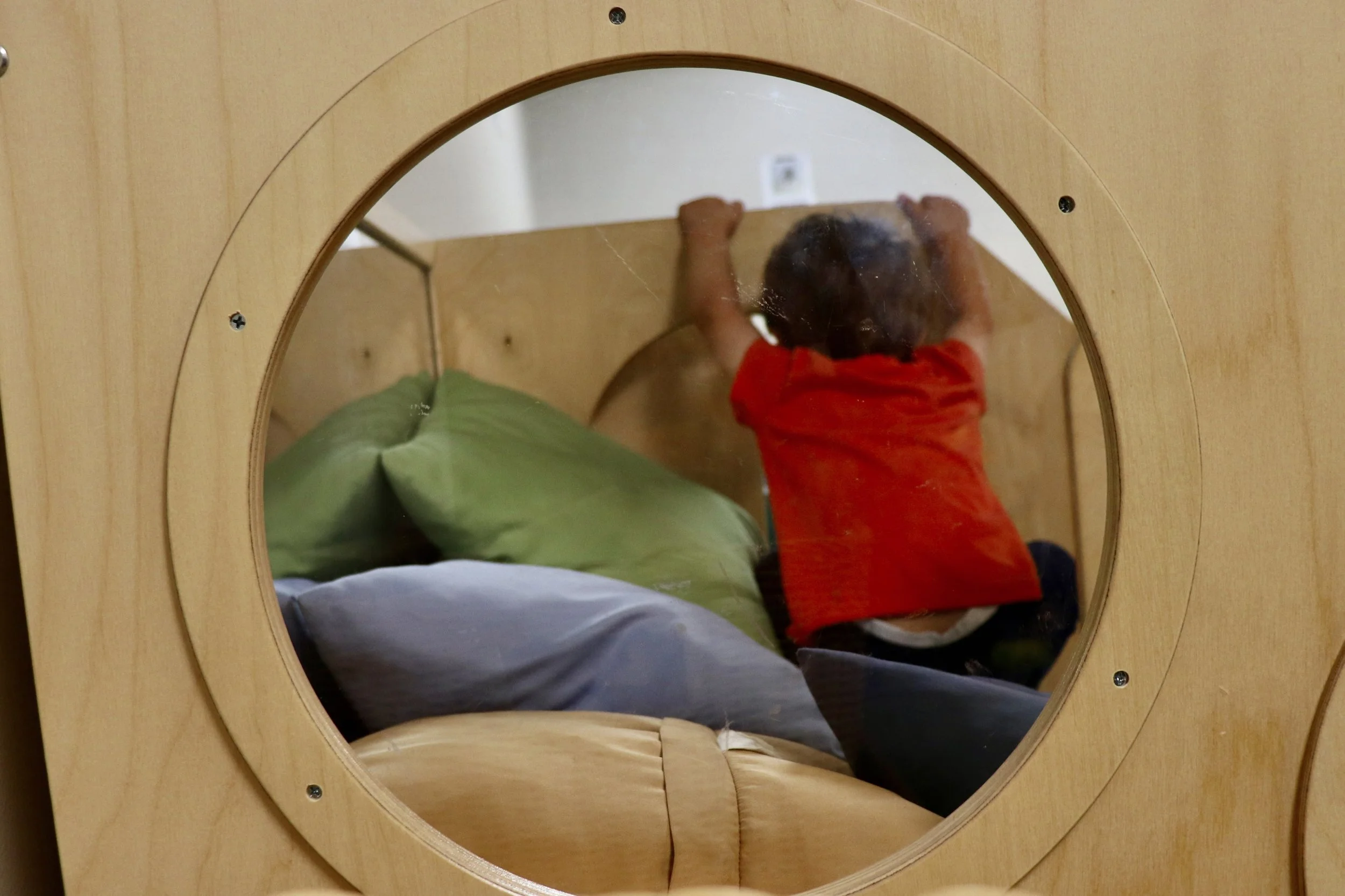 Child in red shirt and dark pants, holding onto a wooden climbing structure, viewed through a circular mirror.