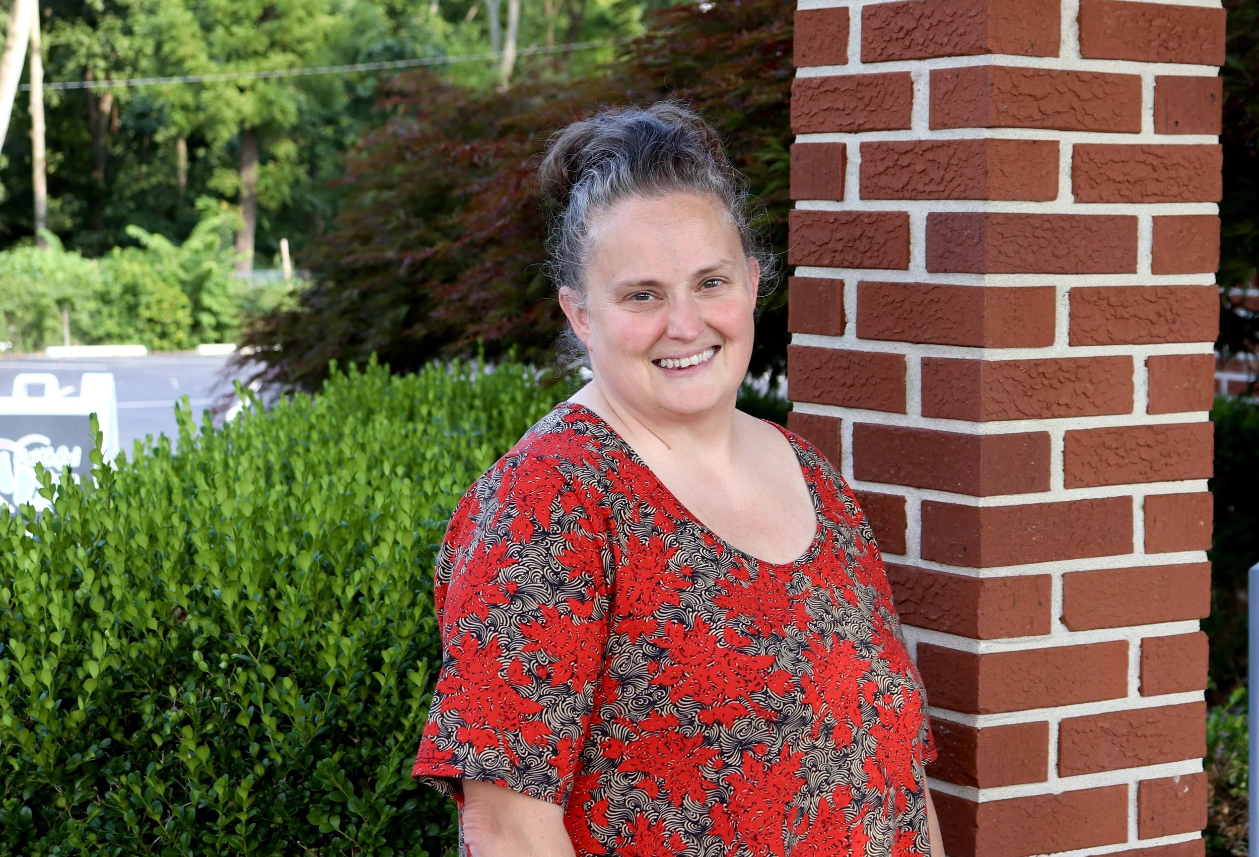 A woman with curly gray hair standing next to a brick pillar, smiling, outdoors with green foliage in the background.