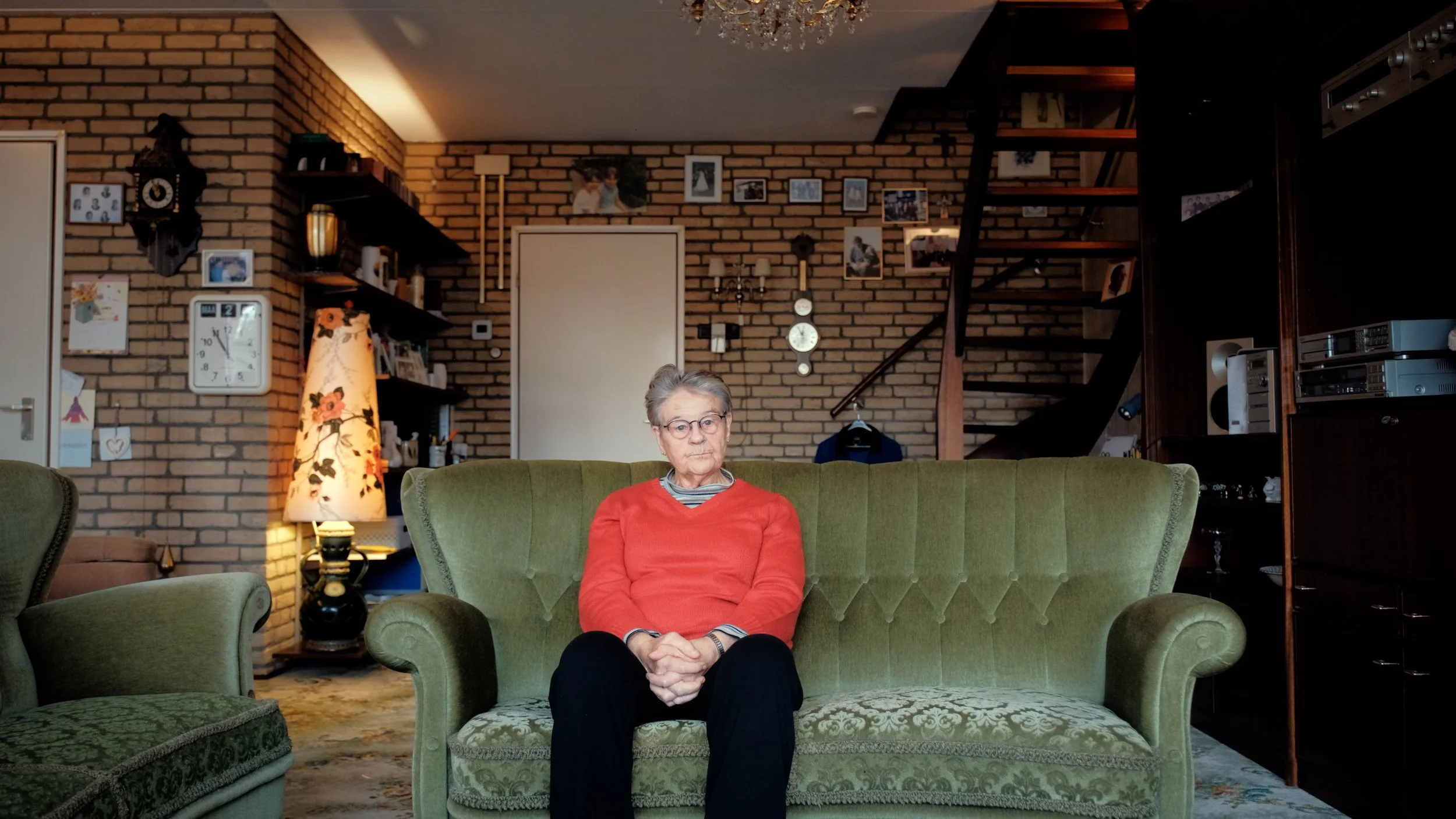 Een oudere vrouw zit op een groene bank in een woonkamertje met bakstenen muren, omringd door meubels en decoraties. Ze kijkt recht vooruit met een neutrale uitdrukking.