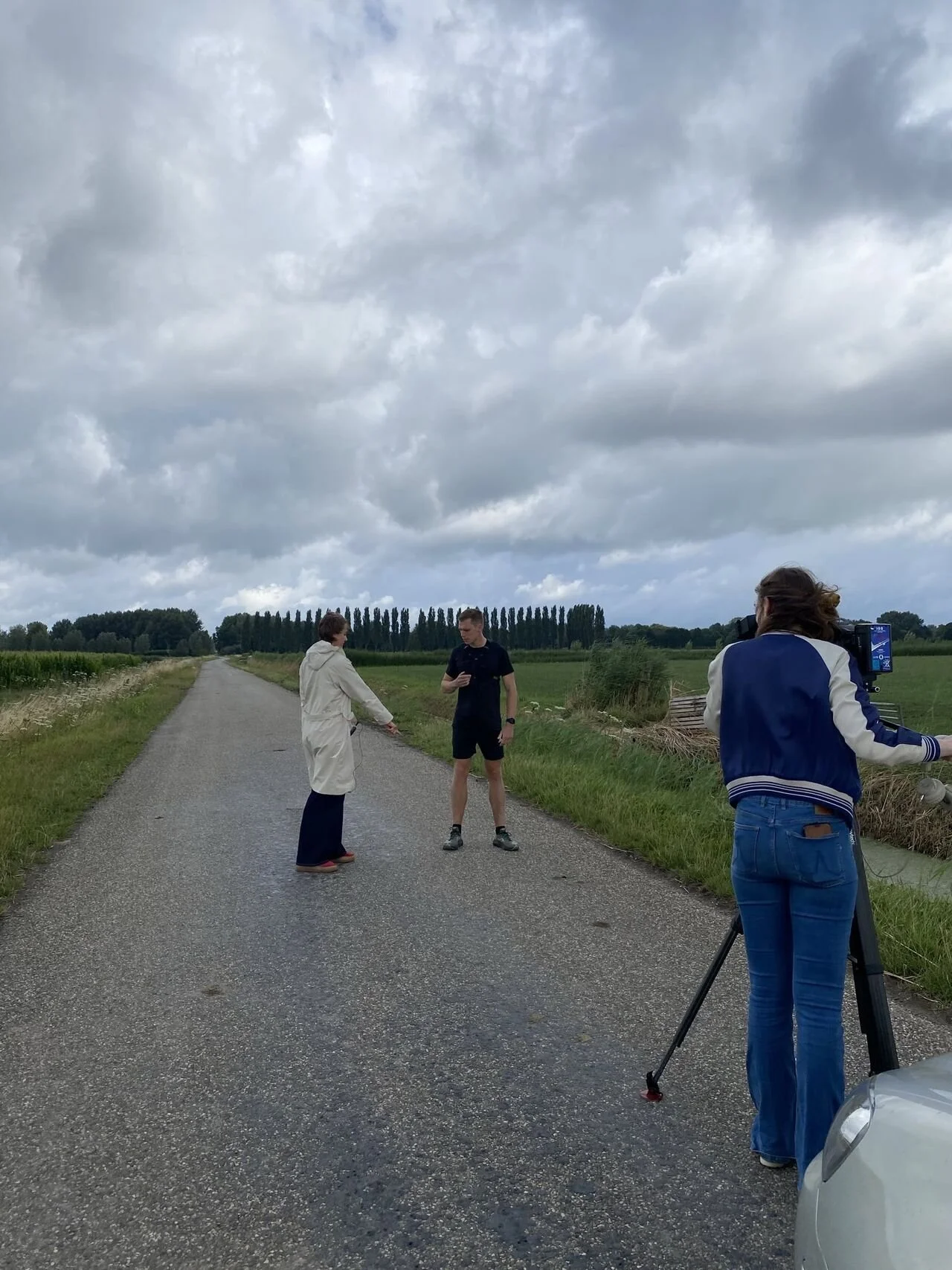 Drietal mensen werken samen op een landweg, met één persoon die foto's maakt met een camera op een statief en twee anderen die voorovergebogen en geïnteresseerd kijken, onder een bewolkte hemel, in een landelijke omgeving.