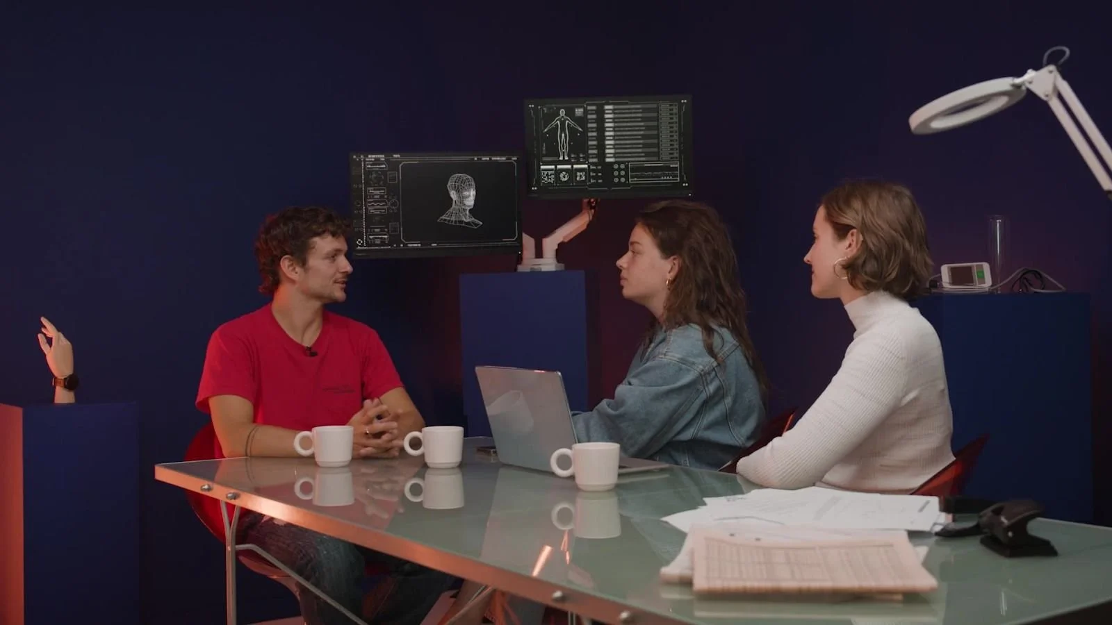 Drie mensen hebben een gesprek in een vergaderruimte met een donkerblauwe muur, een glazen tafel, papieren, een laptop en drie witte mokken. Er zijn twee computerschermen boven hen met medische scans en gegevens.