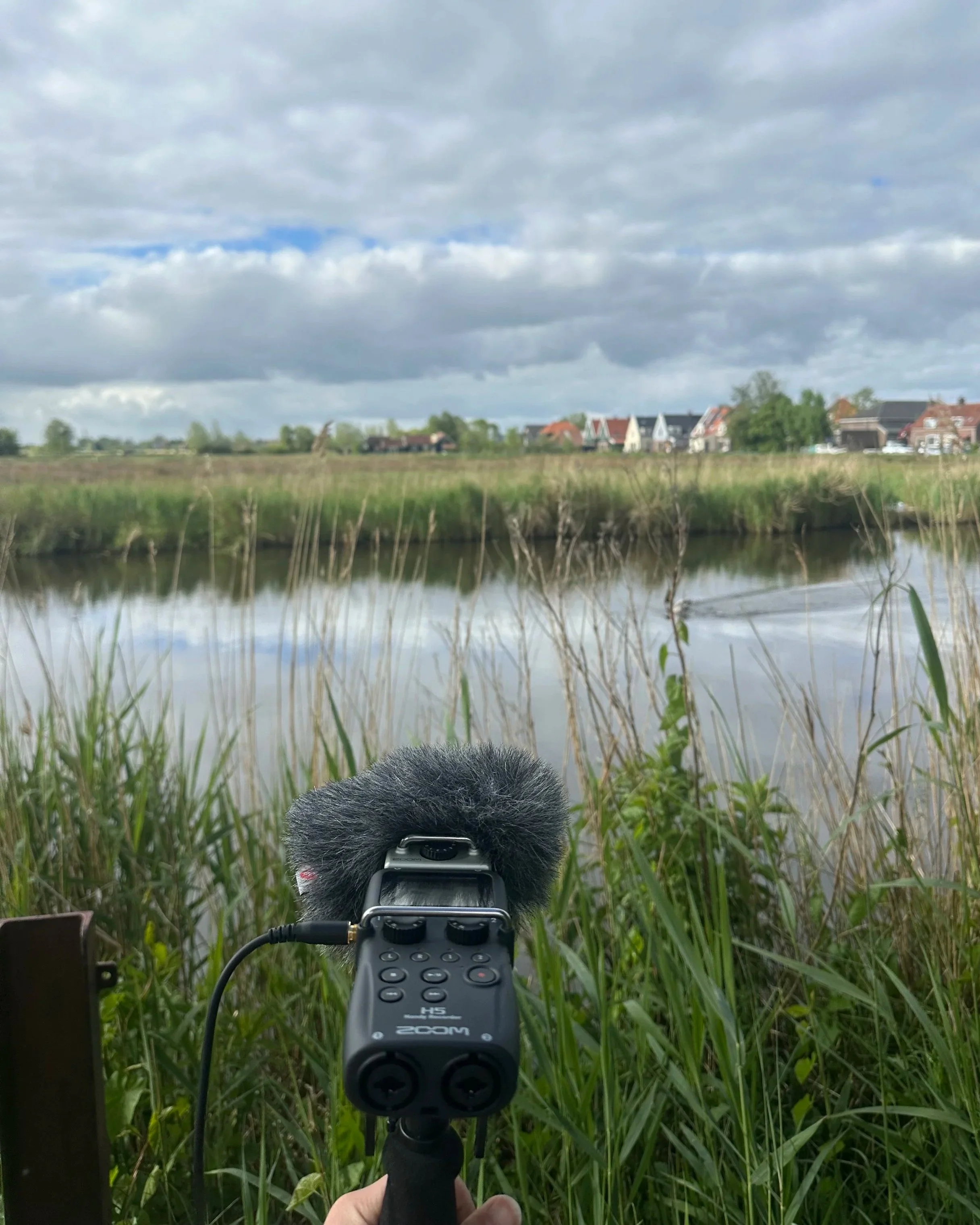 Een camera met een microfoon en een afstandsbediening wordt gebruikt door iemand in een groene grasweide, gericht op een groot landschap met huizen en een waterkant onder een bewolkte hemel.