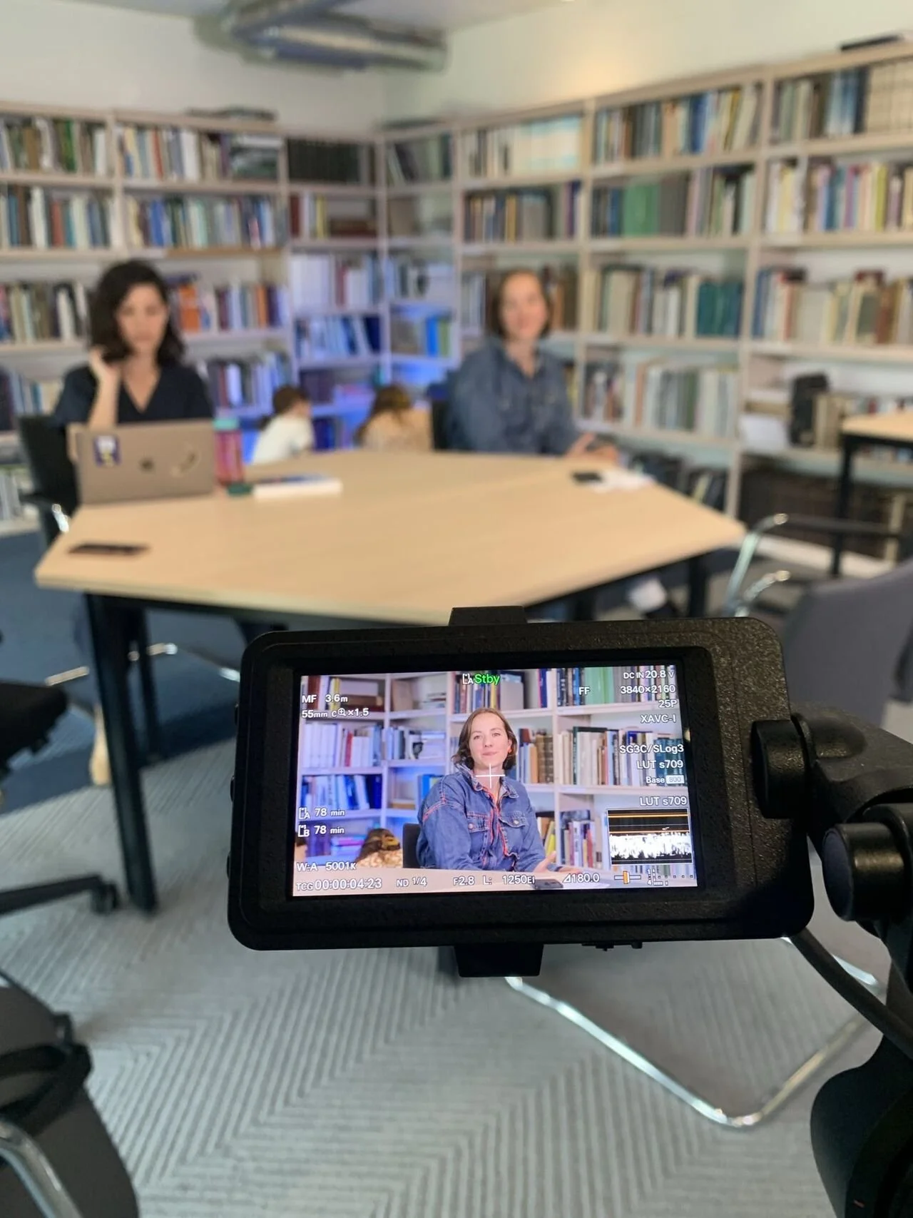 Een filmset of opname studio met een camera gericht op een vrouw in een kamer met boekenplanken. Op scherm is dezelfde vrouw te zien voor de boekenwinkel.