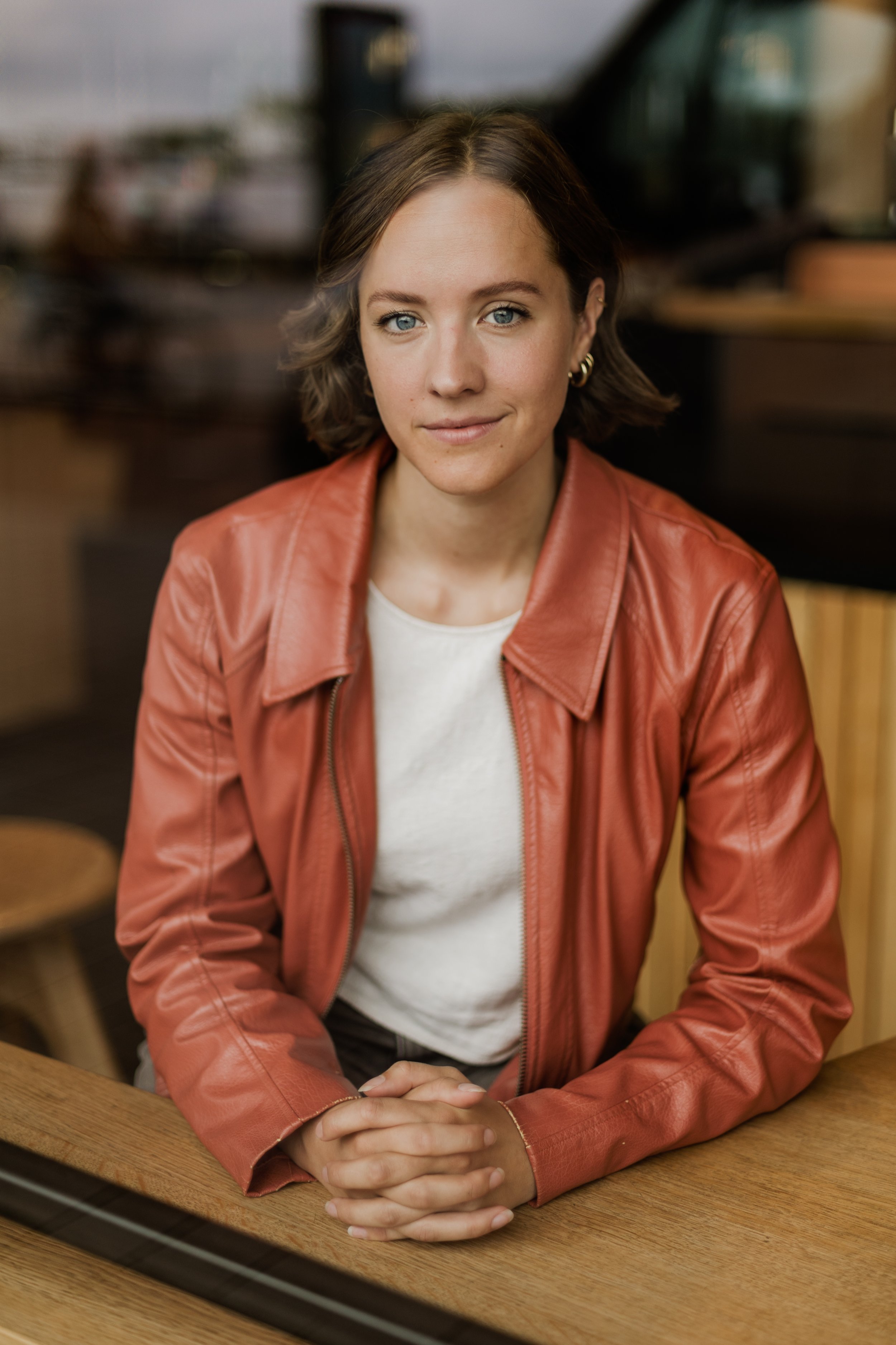 Portret van een jonge vrouw met blauwe ogen, bruin haar, een gouden oorbellen en een oranje leren jack, zittend aan een houten tafel in een café of restaurant.