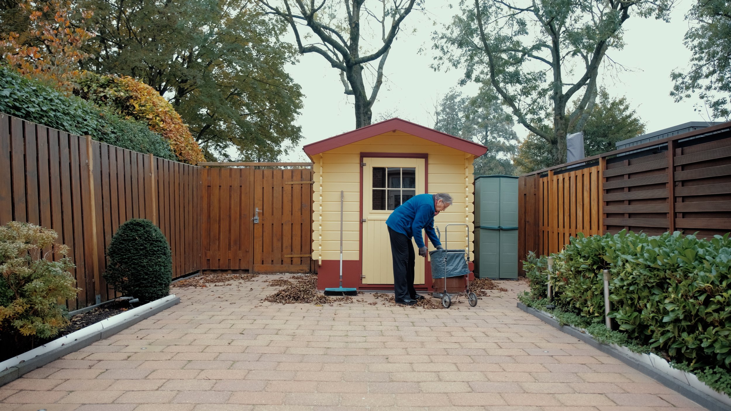 Een man in een blauwe jas veegt het opritje voor een kleine gele en rode tuinkamer in een tuin met planten, bomen en een houten schutting.