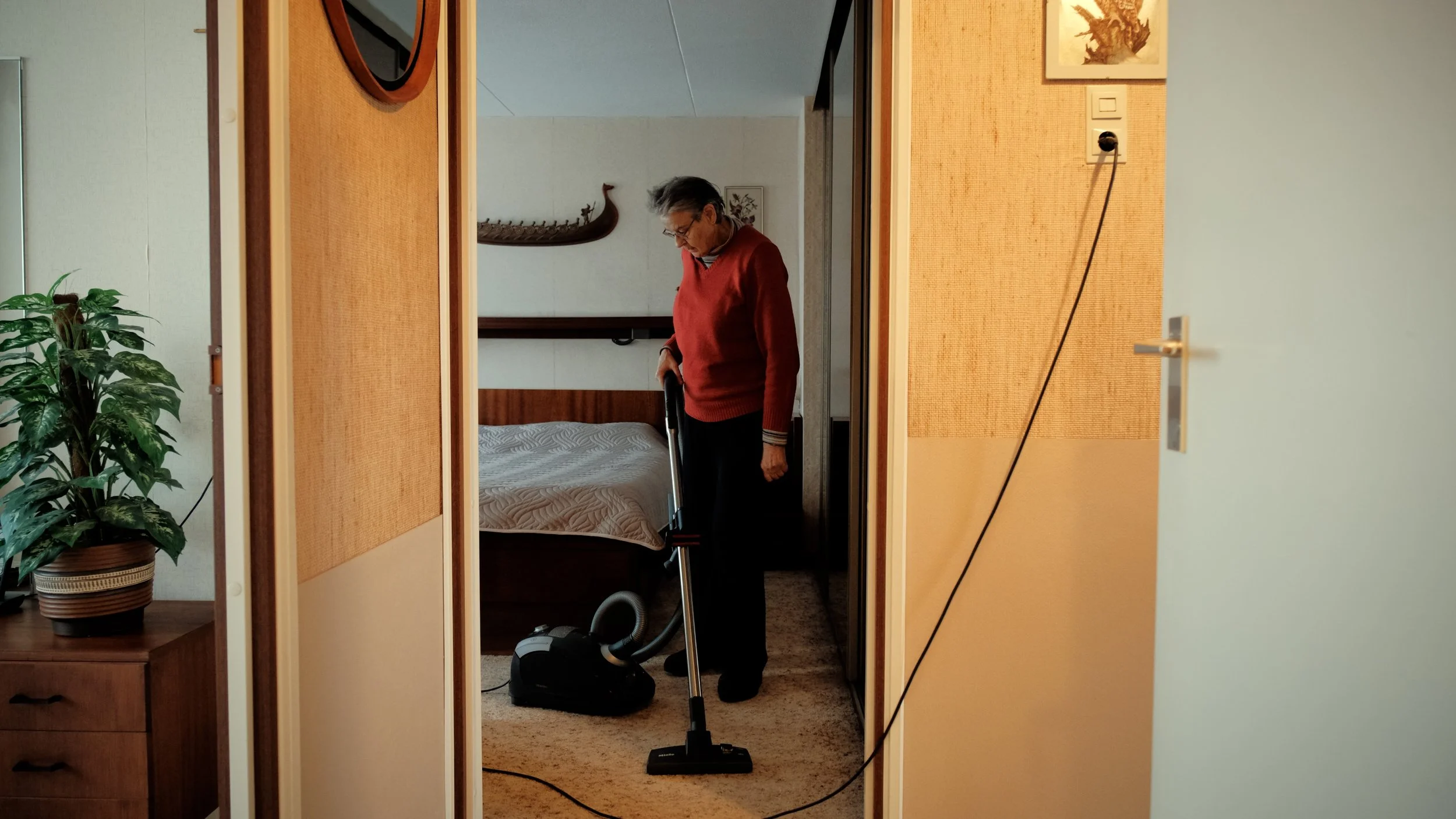 Een oudere man met grijs haar en een rode trui die een stofzuiger gebruikt in een slaapkamer,