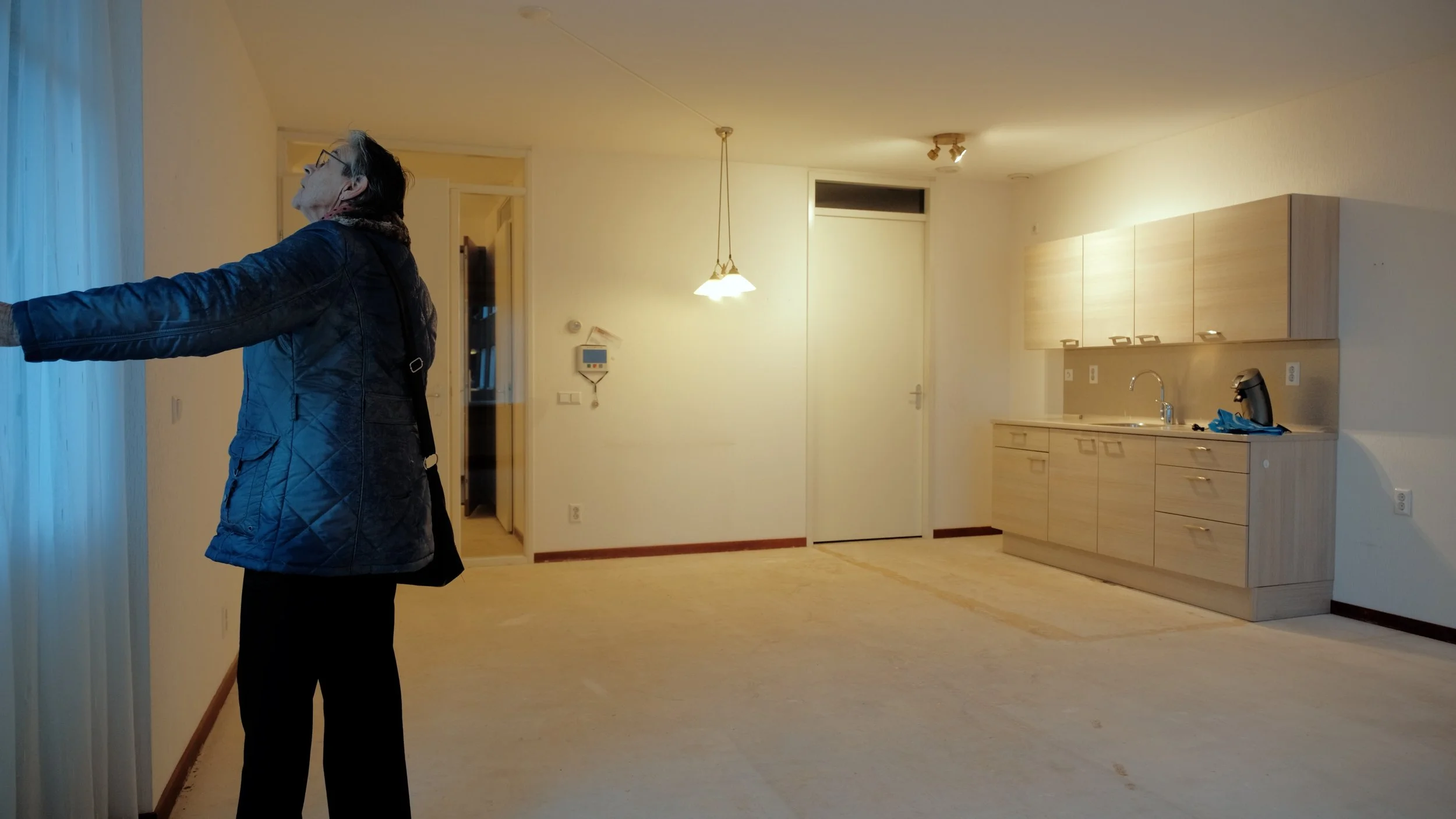 Een vrouw staat binnen in een lege kamer met een keuken, kijkt naar buiten door een raam of deur, met haar arm uitgestrekt.
