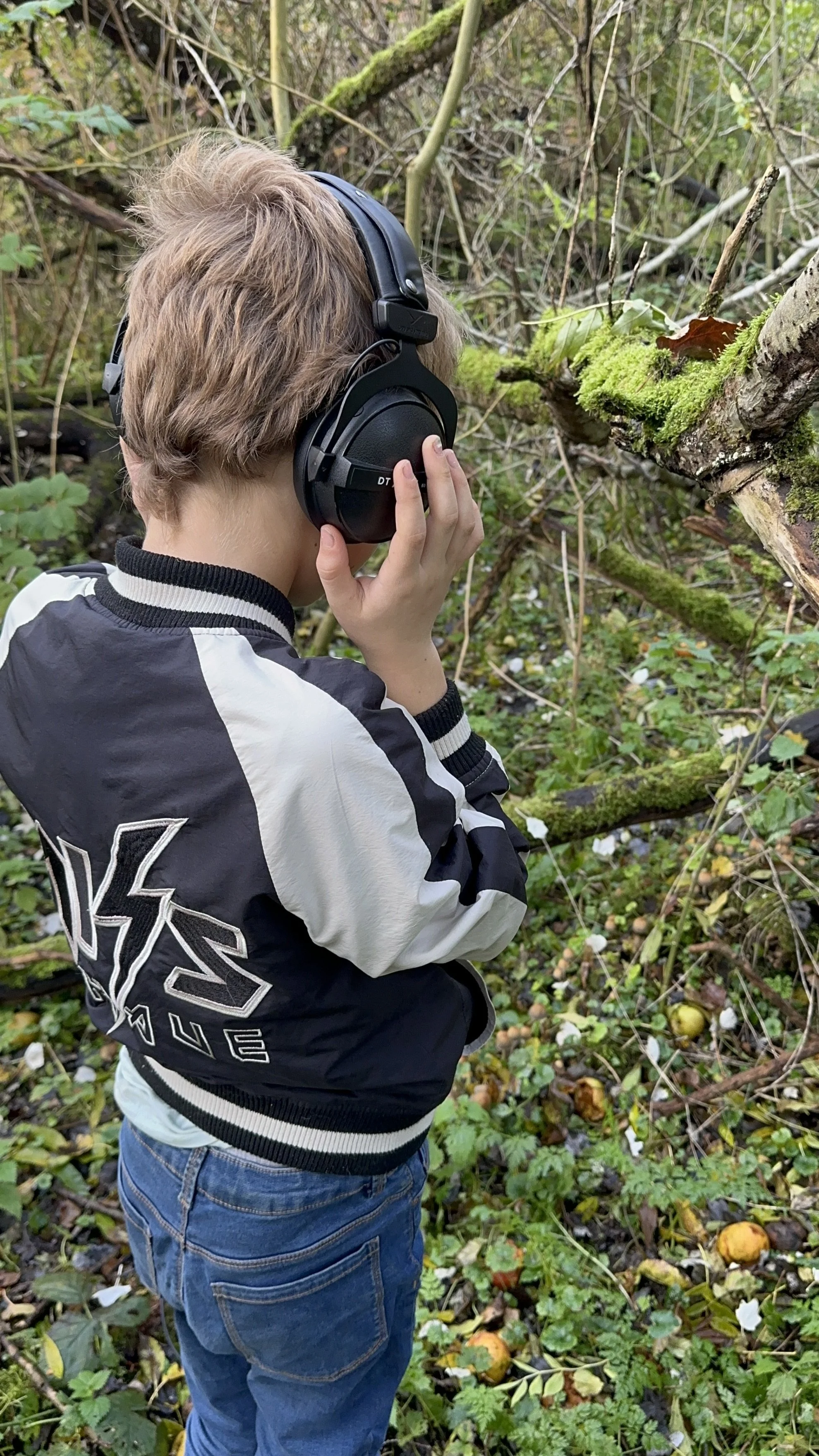Een jongen met schouderlange, lichte schoenen, die in een bos met veel groen en gevallen fruit staat en naar een boom kijkt, terwijl hij een hoofdtelefoon draagt en aan zijn oor houdt.