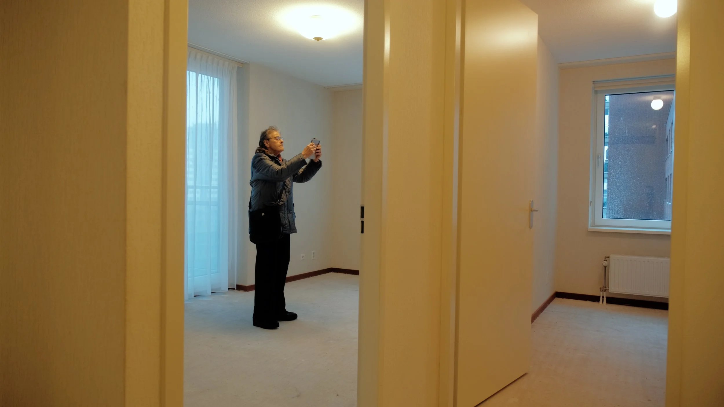 Een vrouwelijkheid die een smartphone vasthoudt in een lege, neutraal ingerichte kamer met grote ramen en gordijnen.
