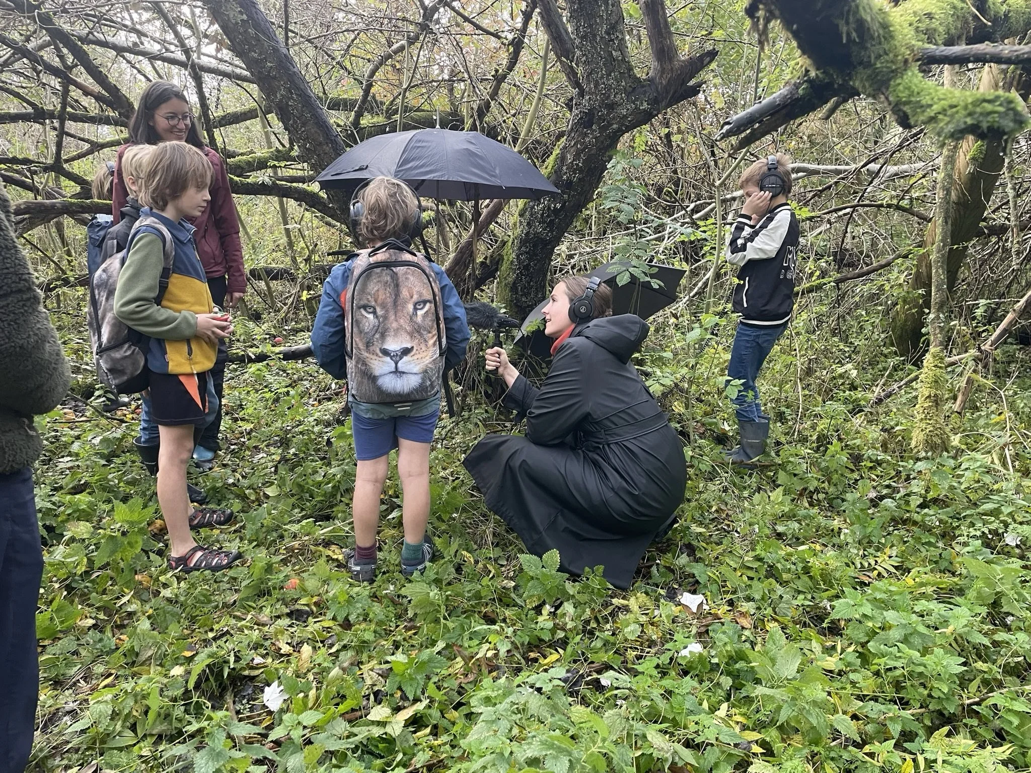 Kinderen en een vrouw in een bos, luisteren naar iemand met een microfoon, enkele kinderen dragen koptelefoons en backpacks, er hangt een paraplu, veel bladeren op de grond.