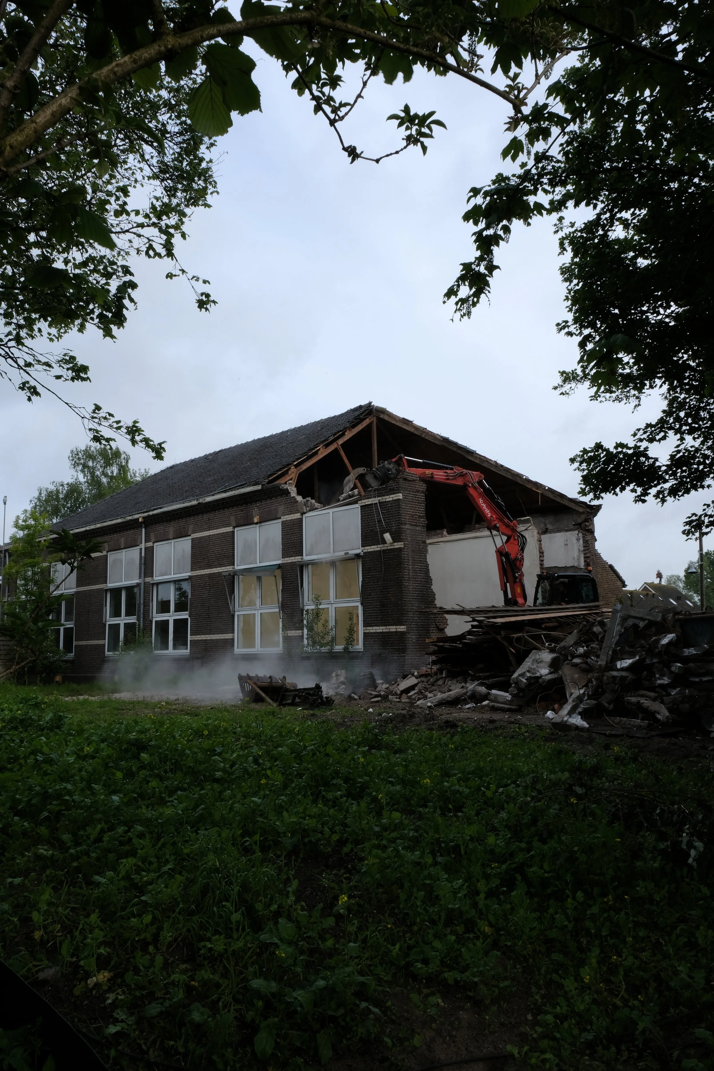 Een huis dat wordt afgebroken met een graafmachine, er ligt puin op de grond, en er wordt rook of stof uit de resten van het gebouw geblazen, omgeven door groen gras en bomen.