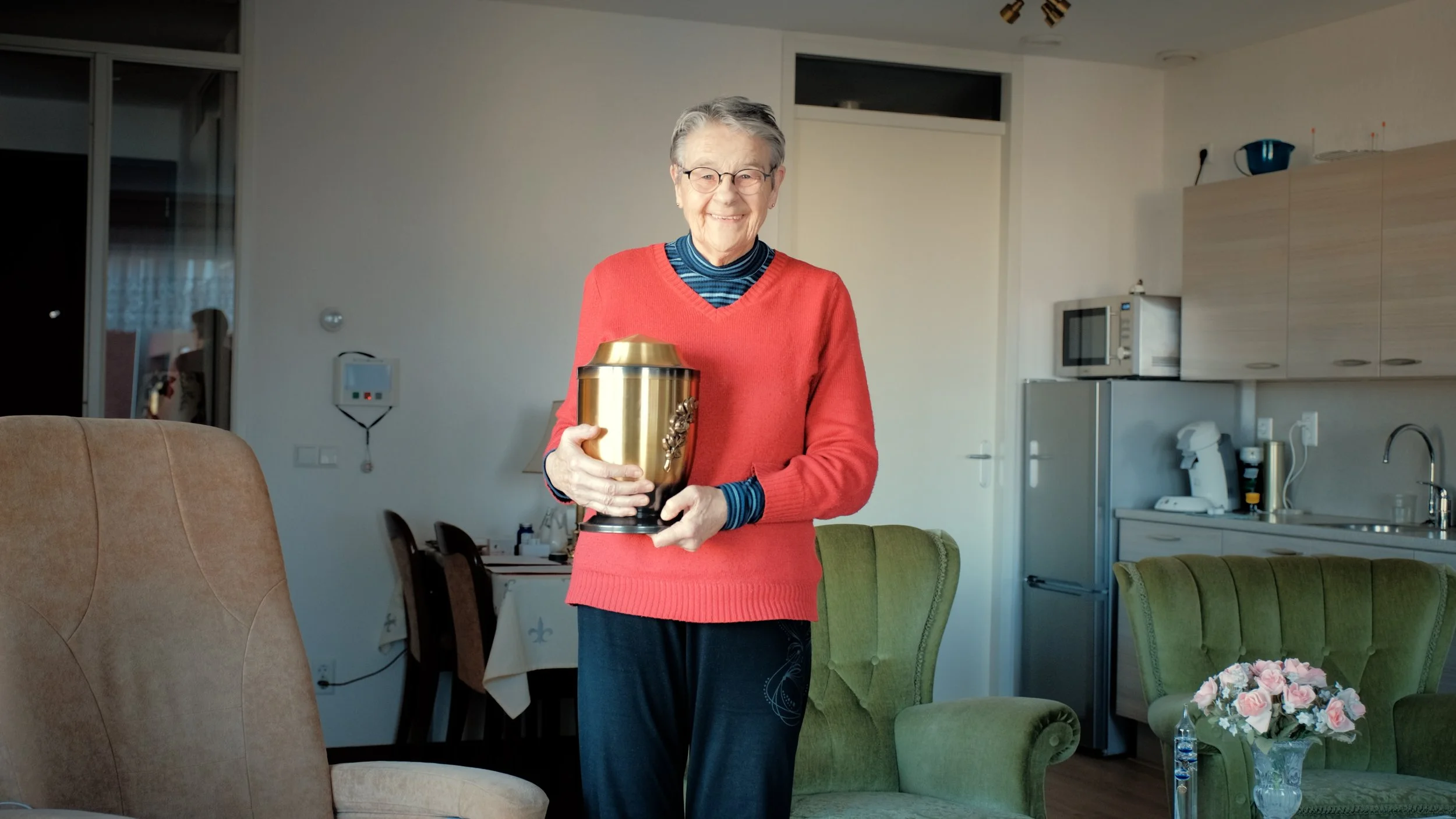 Oudere vrouw met bril en kort grijs haar, glimlachend, vasthoudt een gouden urn met een bloemmotief in de woonkamer.