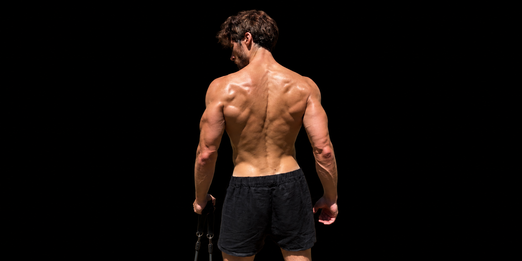 A shirtless man with muscular back and arms, wearing black shorts, standing against a black background, holding a chain in his right hand.