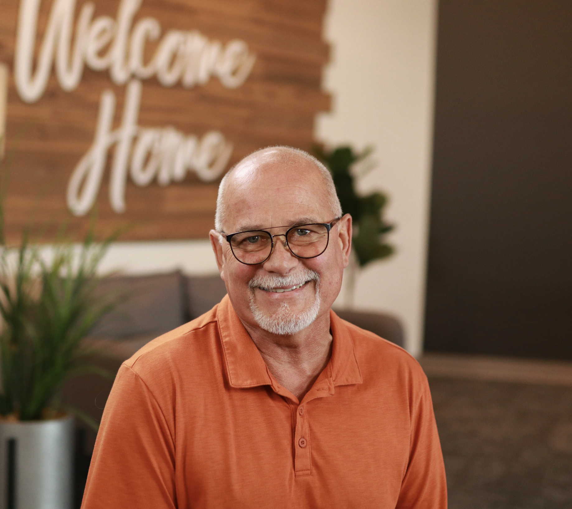 Smiling elderly man with glasses wearing an orange polo shirt indoors with a blurred welcome sign and plant in the background.
