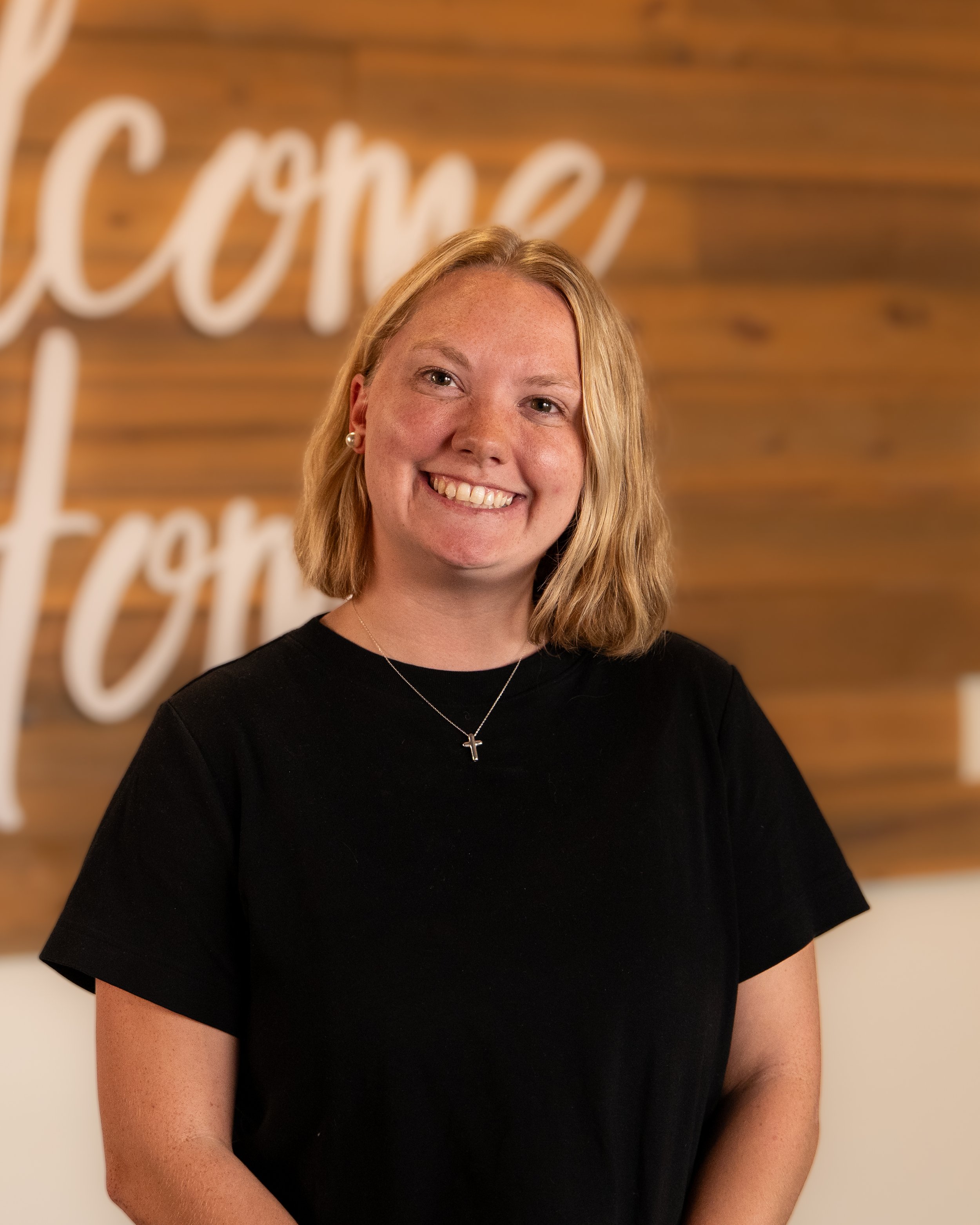 Smiling blonde woman with curly hair wearing a gray sweater, standing in front of a wooden wall with white cursive writing.
