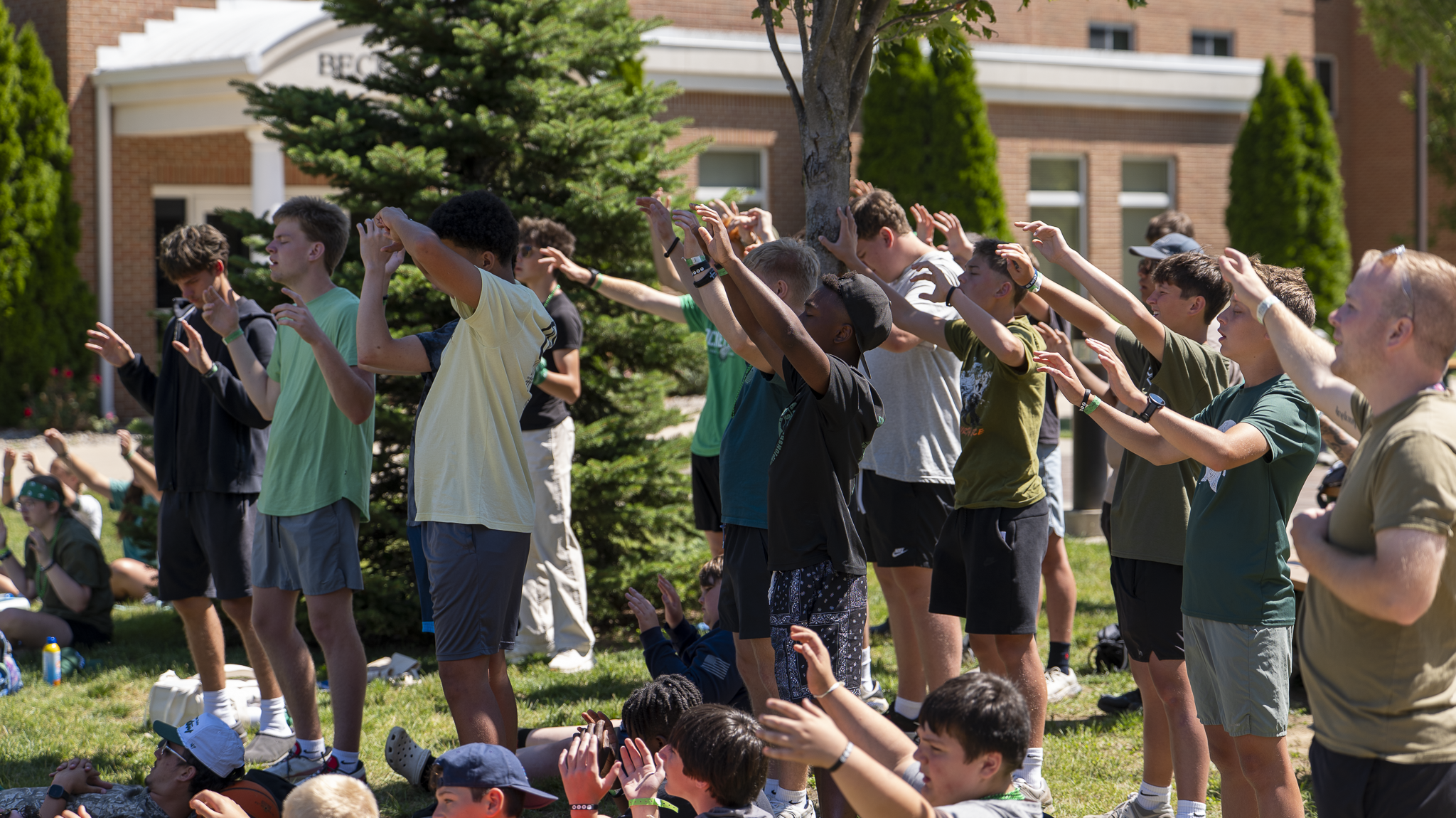 A diverse group of young people participating in an outdoor yoga or meditation class, standing with arms raised, in front of a brick building with trees around.