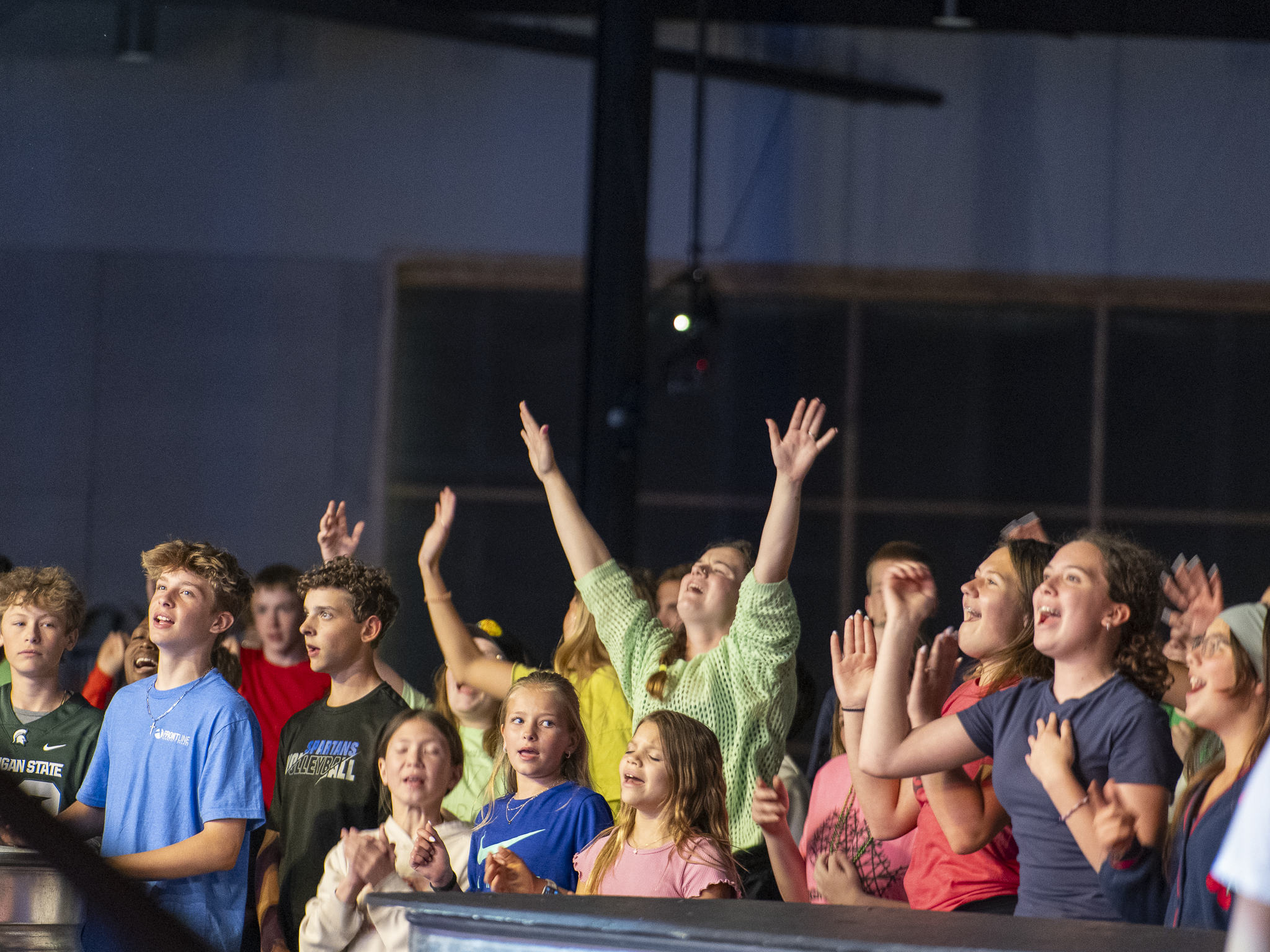A group of diverse children and teenagers at a concert or auditorium, raising their hands, clapping, and enjoying the event, with a dark background and visible stage lighting.
