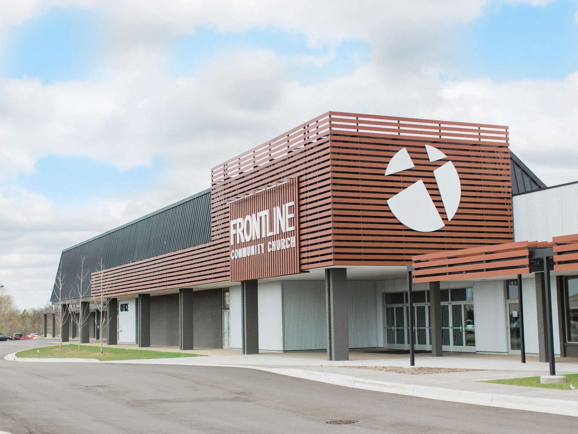 Exterior view of a modern community church building with a sign reading "Frontline Community Church," featuring a large logo of white shapes forming a cross. The building has a combination of dark gray and wood-paneled exterior with large windows and a parking lot in front.