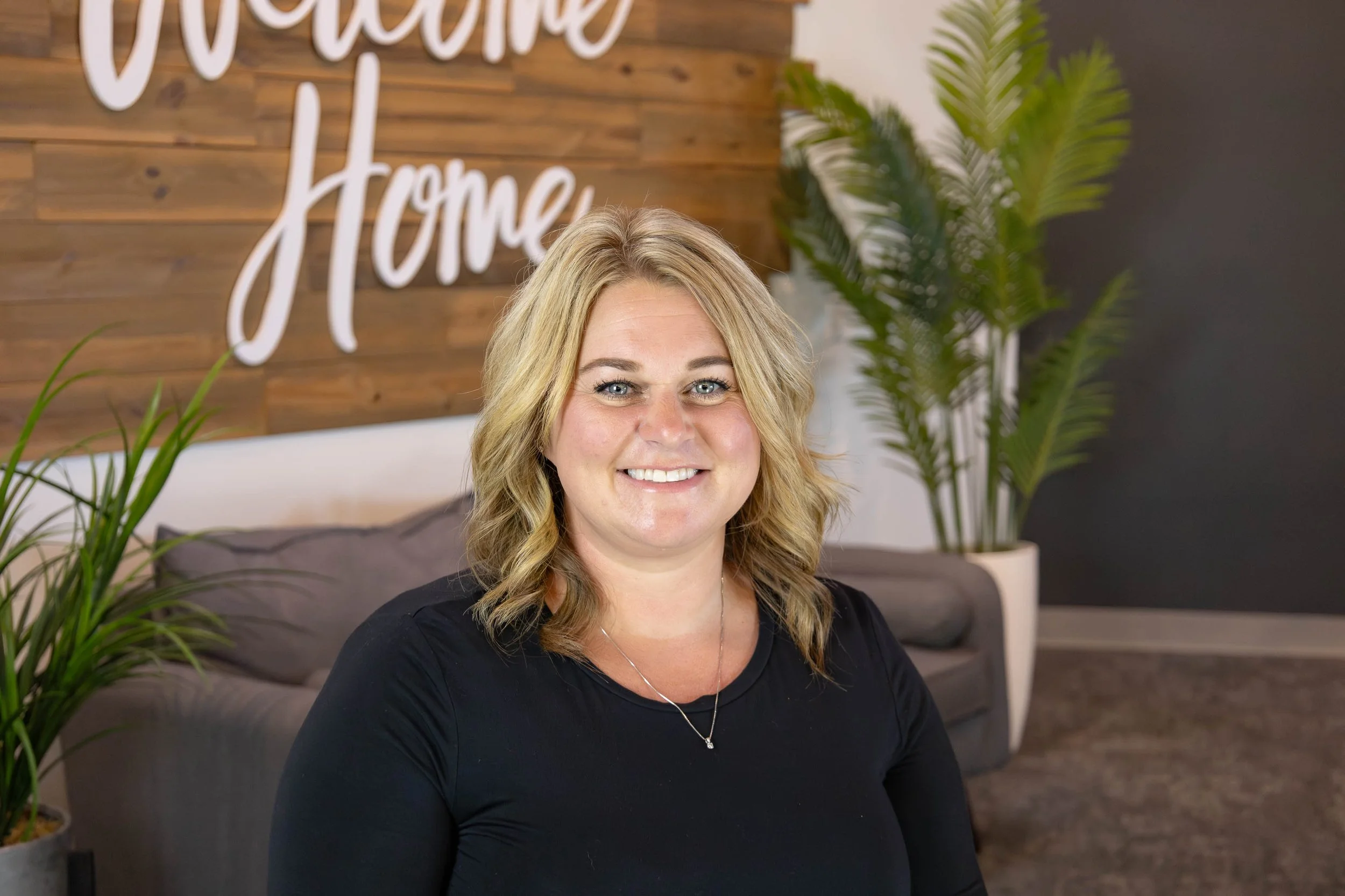 A smiling woman with blonde hair sitting in a modern indoor space with green plants and a wooden wall behind her that says 'Welcome Home'.