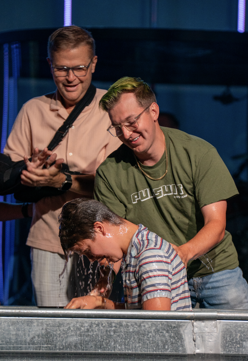A boy getting baptized in a church or religious venue, with two men assisting him in the baptism process.