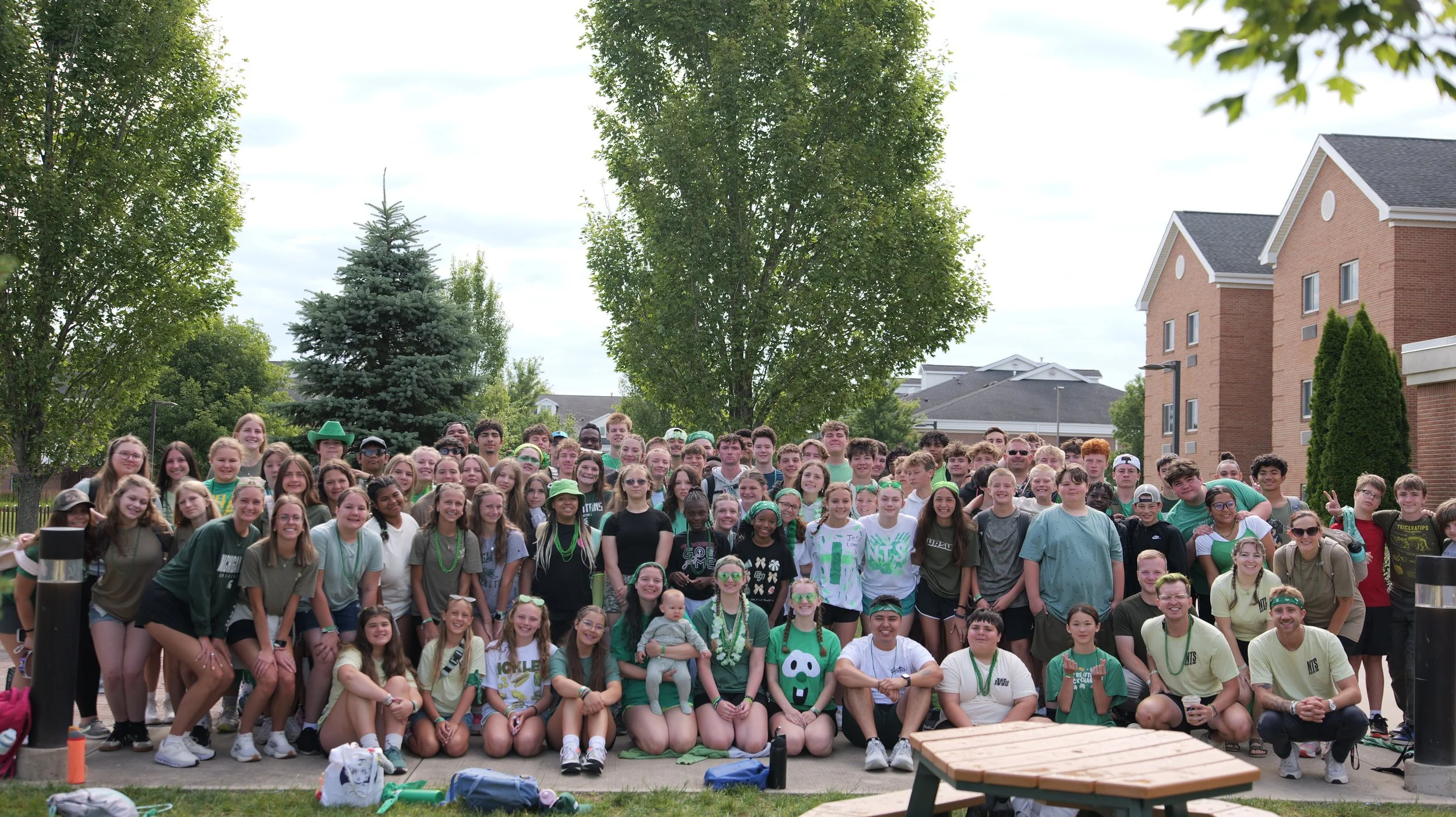A large group of young students and a few adults gathered outdoors in front of trees and residential buildings, smiling for a group photo during a sunny day.