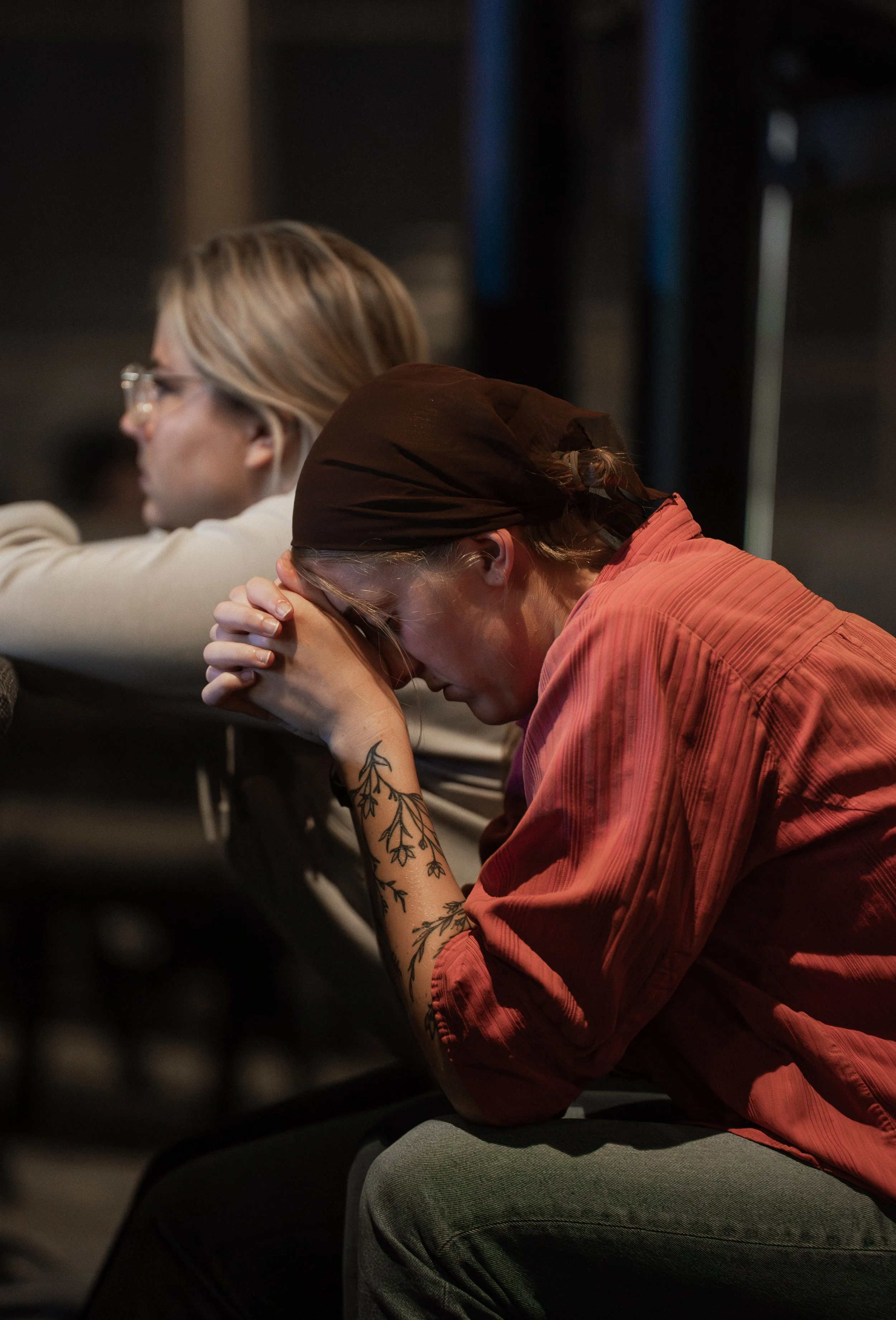 Two women sitting with heads bowed and hands clasped, appearing to be praying in a dimly lit room.