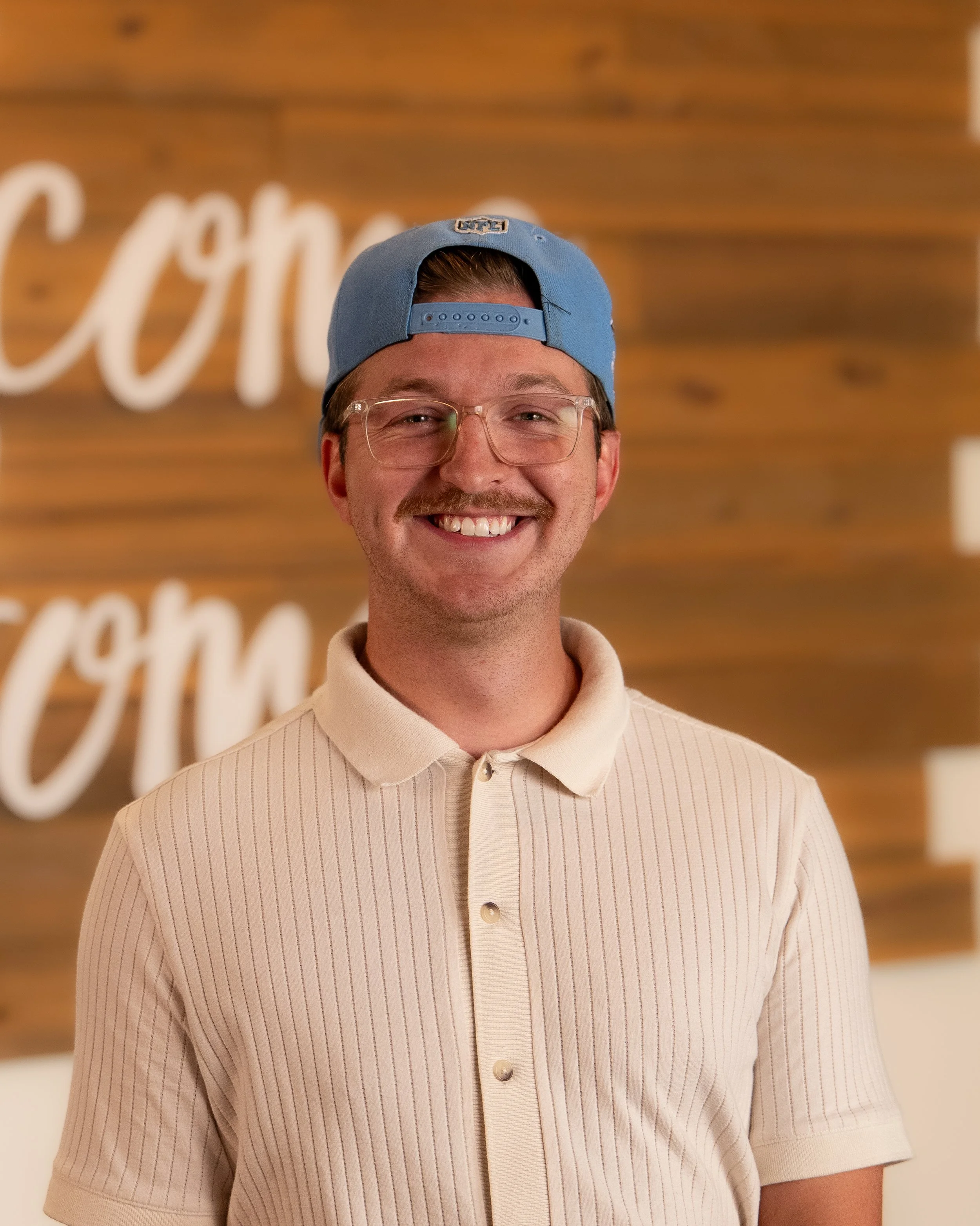 A smiling young man with glasses and styled hair, wearing a white shirt, standing indoors with a wooden wall, decorative sign, and green plant in the background.
