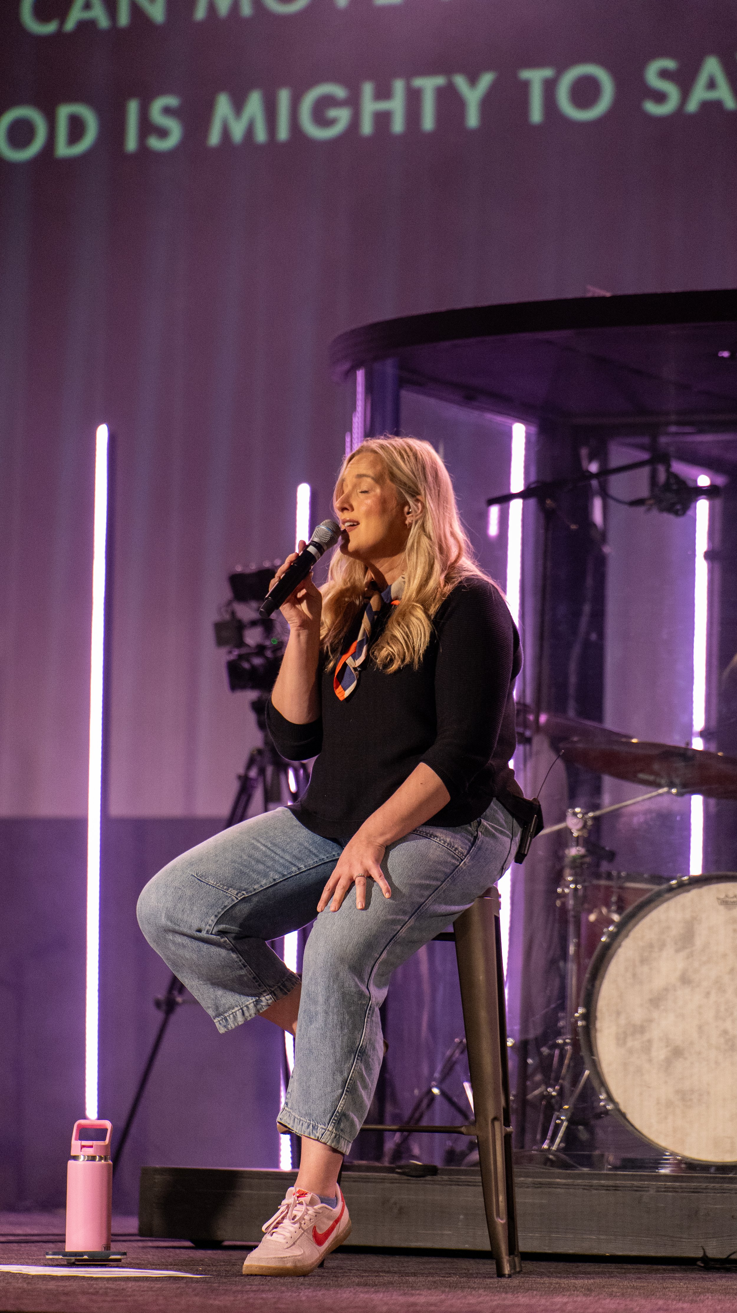 A woman singing into a microphone on stage, sitting on a stool. She has long blonde hair, is wearing a black top, jeans, and pink Nike sneakers. Purple stage lighting and a drum set are visible in the background.