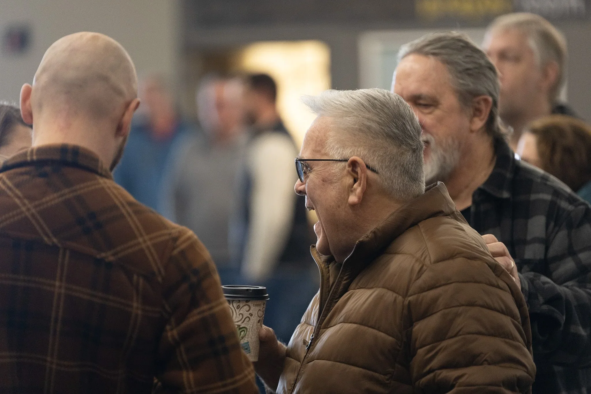 A group of middle-aged people at a social gathering, engaging in conversation, with some holding coffee cups. Focus on a man with gray hair and a woman with glasses sharing a moment.