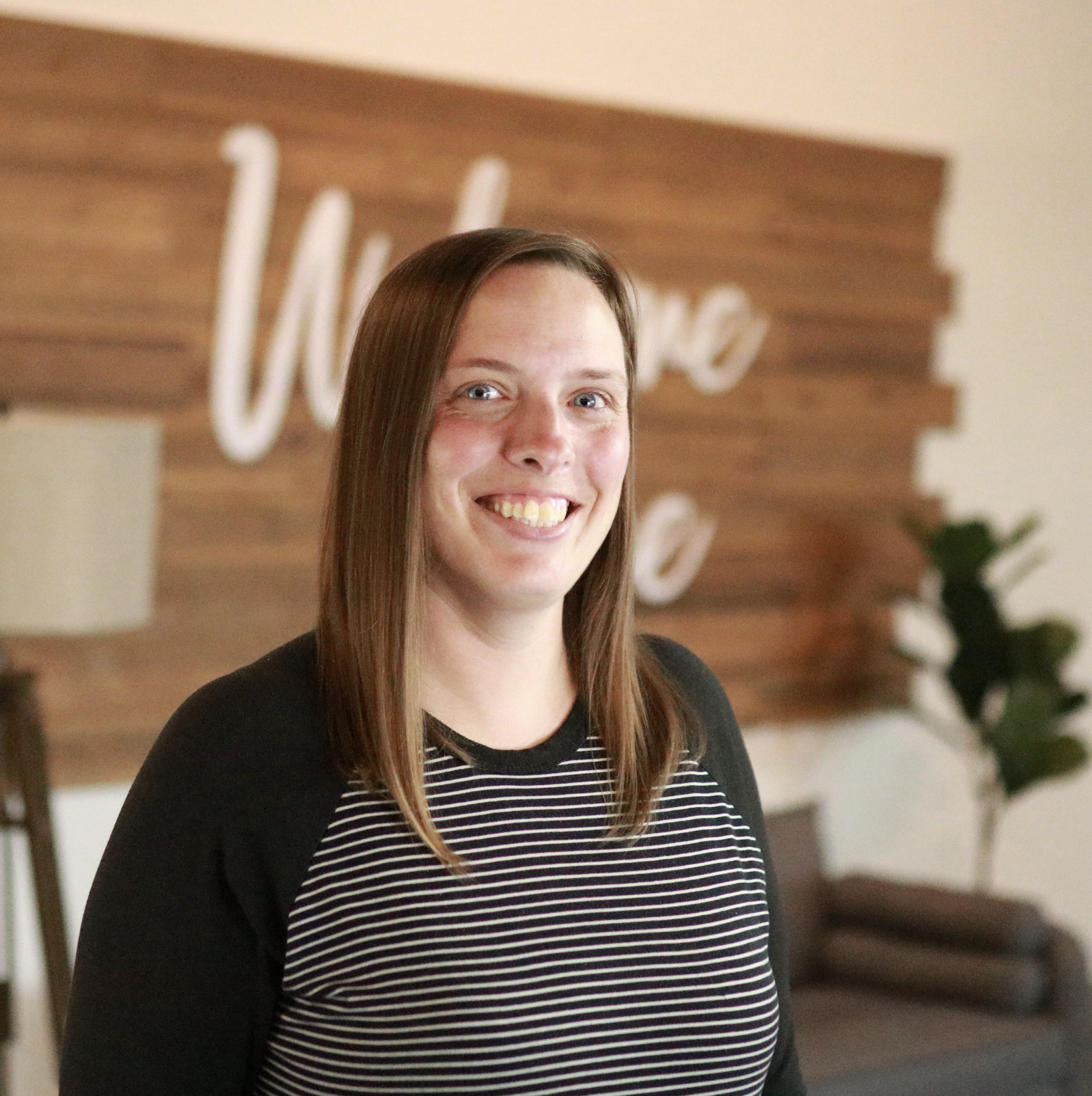 A woman with long brown hair, smiling, wearing a black and white striped shirt and a black cardigan, standing in a room with wooden wall decor and some plants in the background.