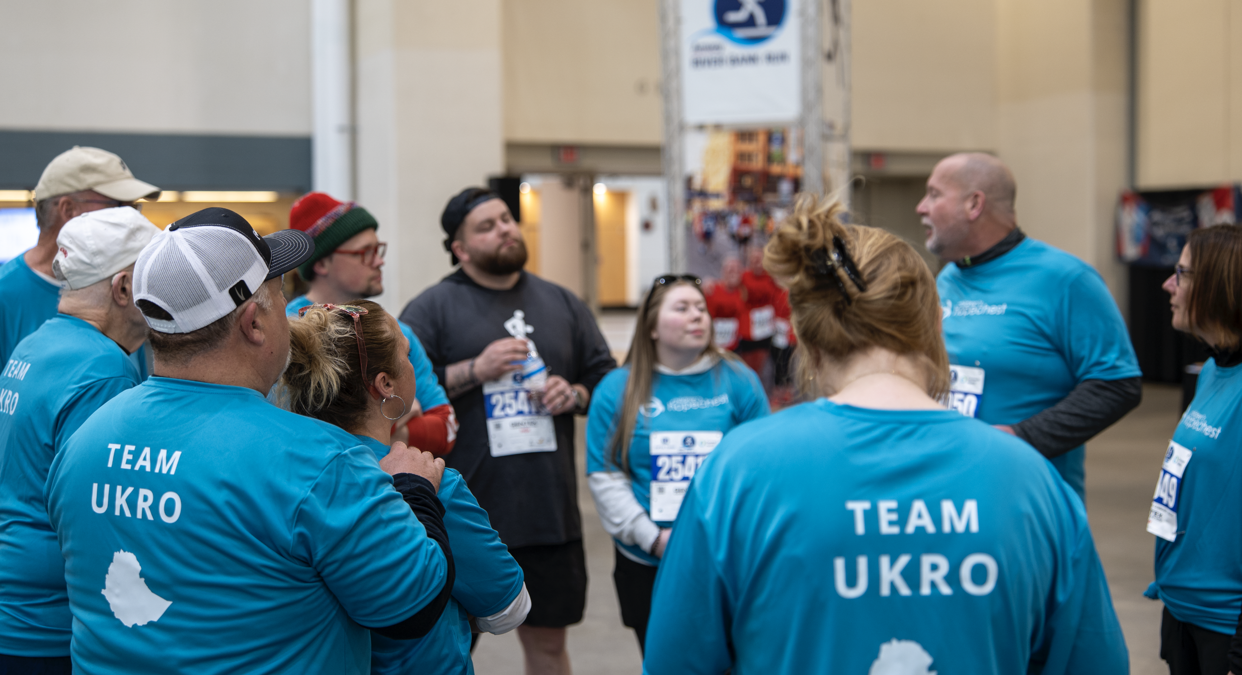 A group of people wearing blue shirts with 'TEAM UKRO' on the back gathered together indoors, some with race bibs, possibly at a marathon or charity event, engaging in a discussion or briefing.