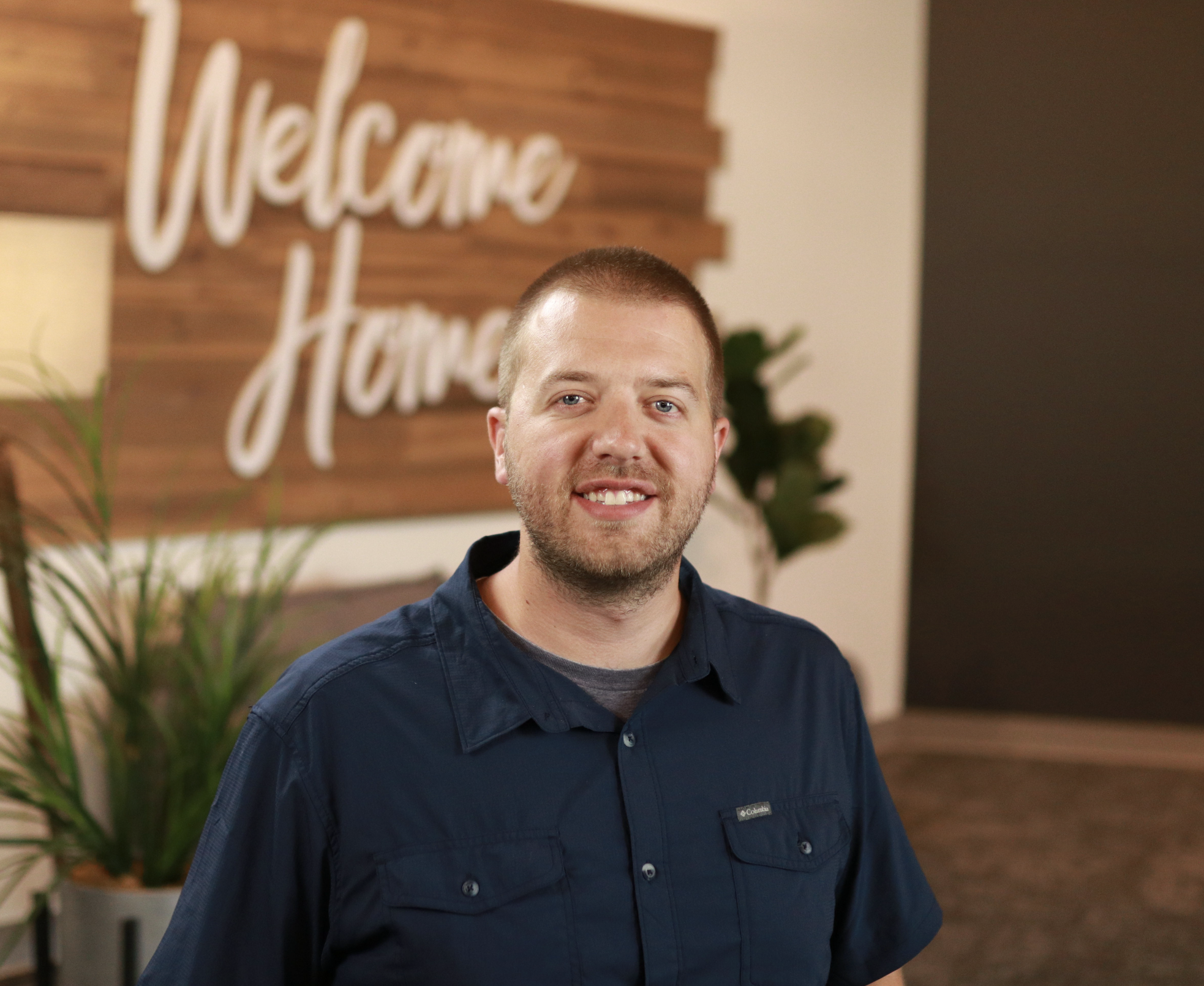 A smiling man with short brown hair and a beard, wearing a navy blue Columbia jacket, standing indoors in front of a wooden wall with a blurred background that includes a large sign saying 'Welcome' and some greenery.