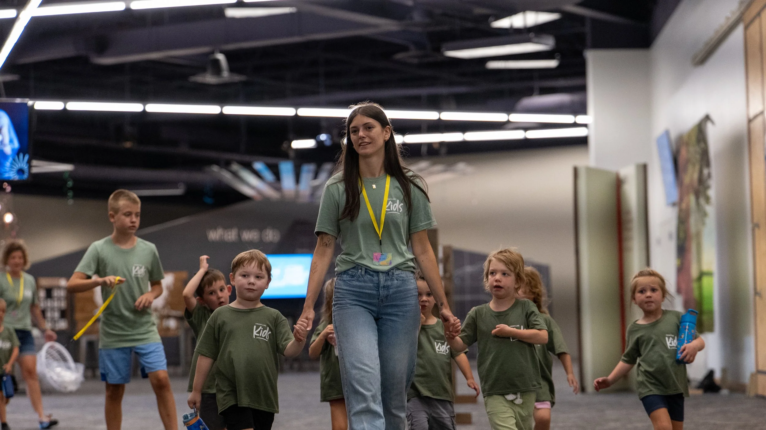 A young woman with long dark hair, wearing a green t-shirt and jeans, leading a group of young children in a children’s activity or camp indoors. The children are also wearing matching green shirts and are holding hands or water bottles.