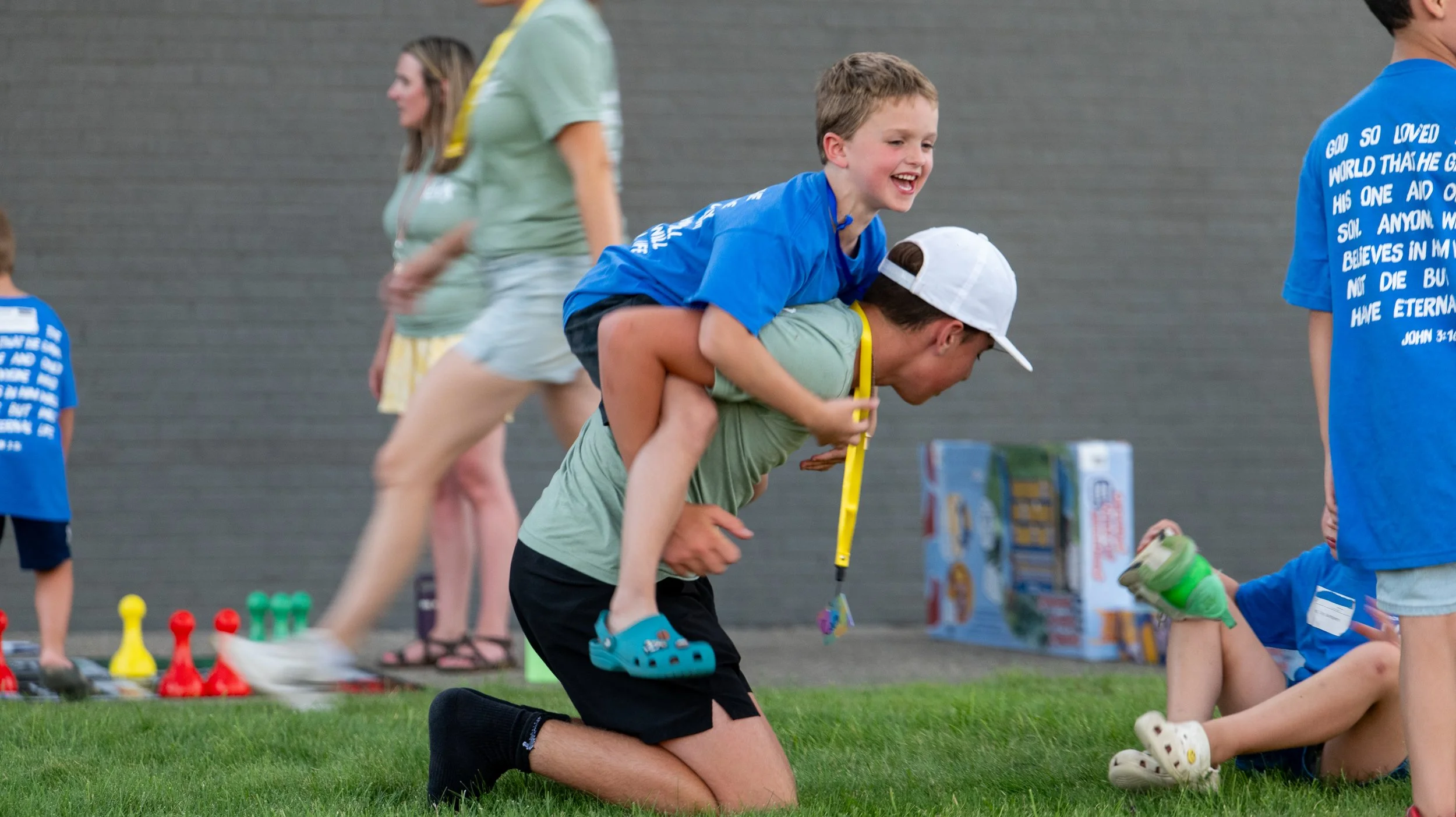 Children playing a game outdoors, with one child on another's back, smiling, near toys and cones on grass, in front of a brick wall.