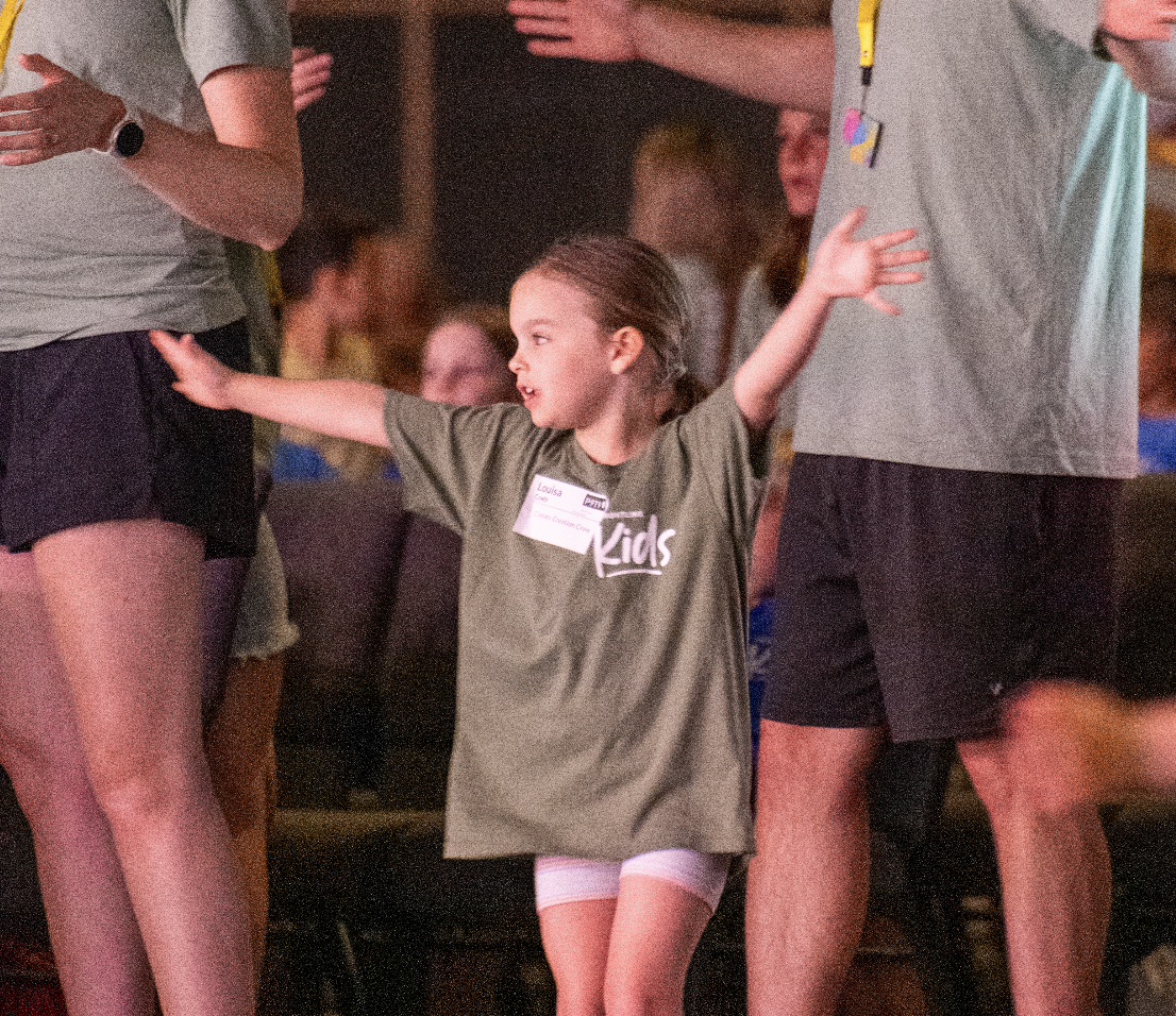 A young girl standing with arms outstretched in a crowded indoor setting, wearing a green kids t-shirt and white shorts, surrounded by adults.