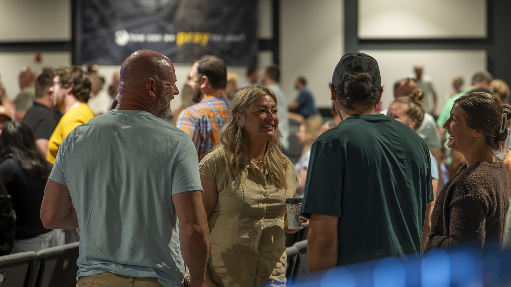 A group of people socializing at an indoor event. Men and women are engaged in conversation, some holding cups. The background shows more attendees and a banner.