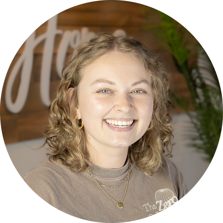 Young woman with curly blonde hair smiling, wearing gold jewelry, in front of a wooden wall with a decorative sign and green plants.
