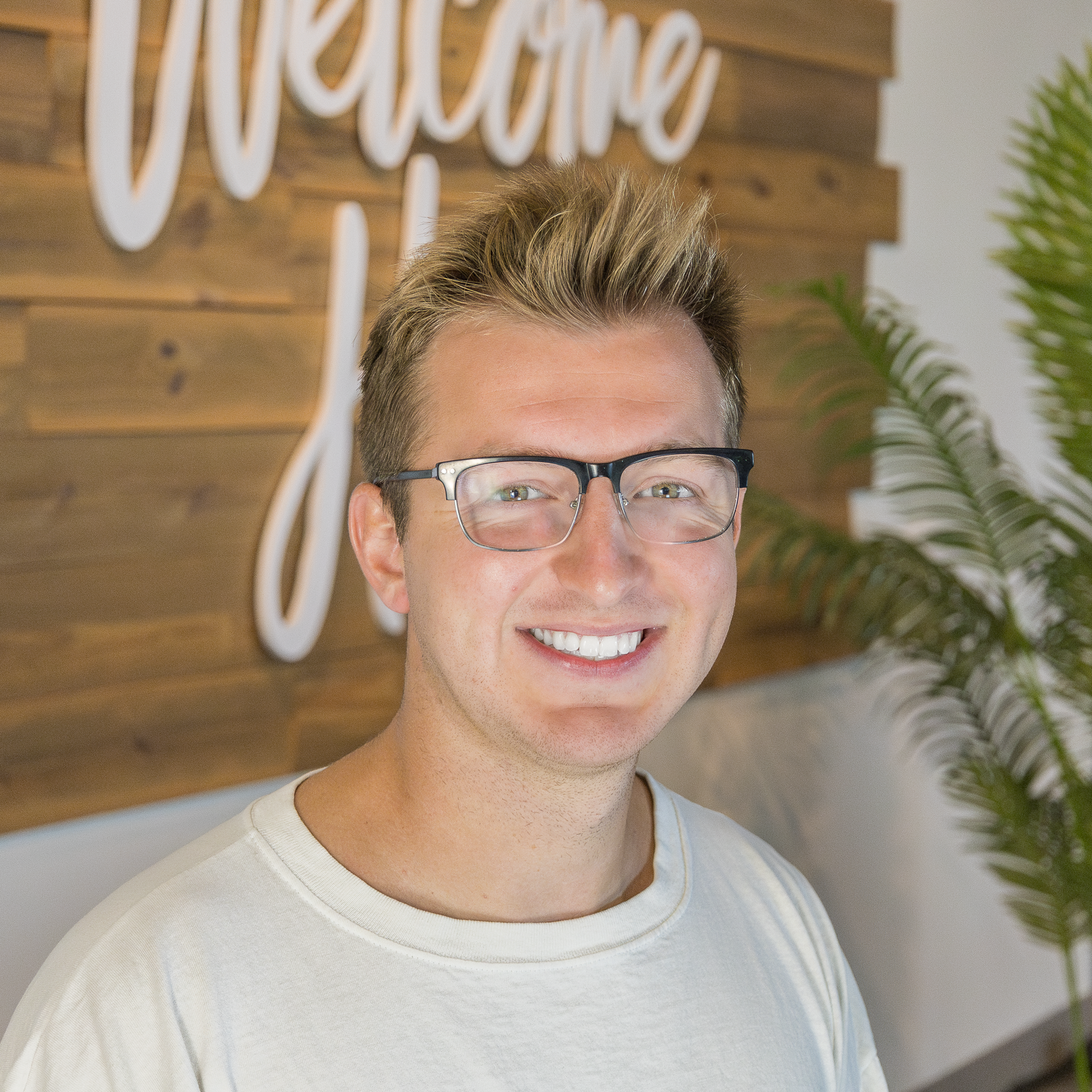 A young man with styled brown hair, wearing glasses and a white t-shirt, smiling in front of a wooden wall with white cursive lettering and a leafy plant.