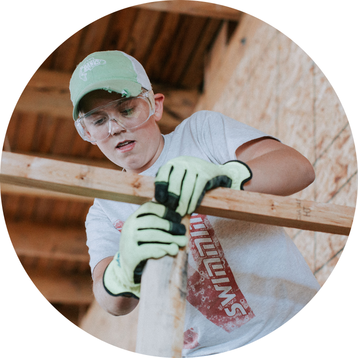 Young man wearing safety goggles, a green cap, a gray T-shirt, and yellow work gloves, working with wood in a carpentry or construction setting.