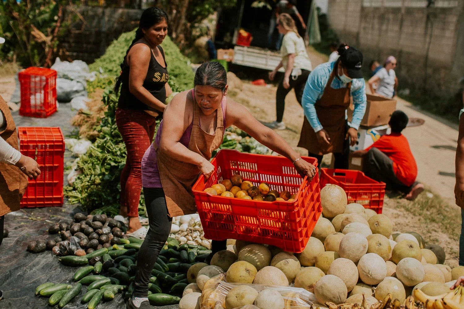 Women shopping for fruits and vegetables at an outdoor market, with produce like melons, cucumbers, and avocados on the ground.