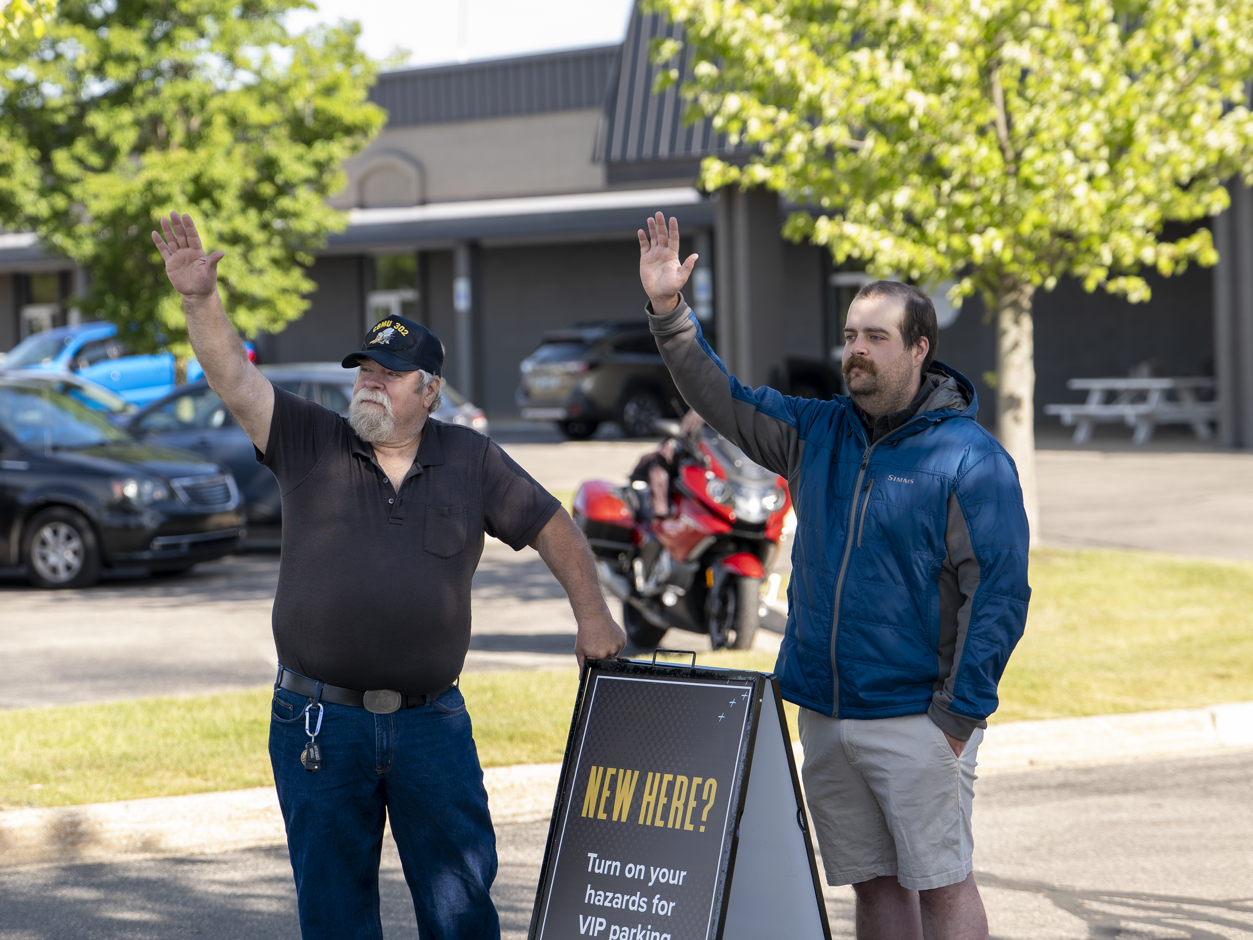 Two men waving in front of a parking lot with trees and cars, next to a sign that reads 'NEW HERE? Turn on your hazards for VIP parking'.