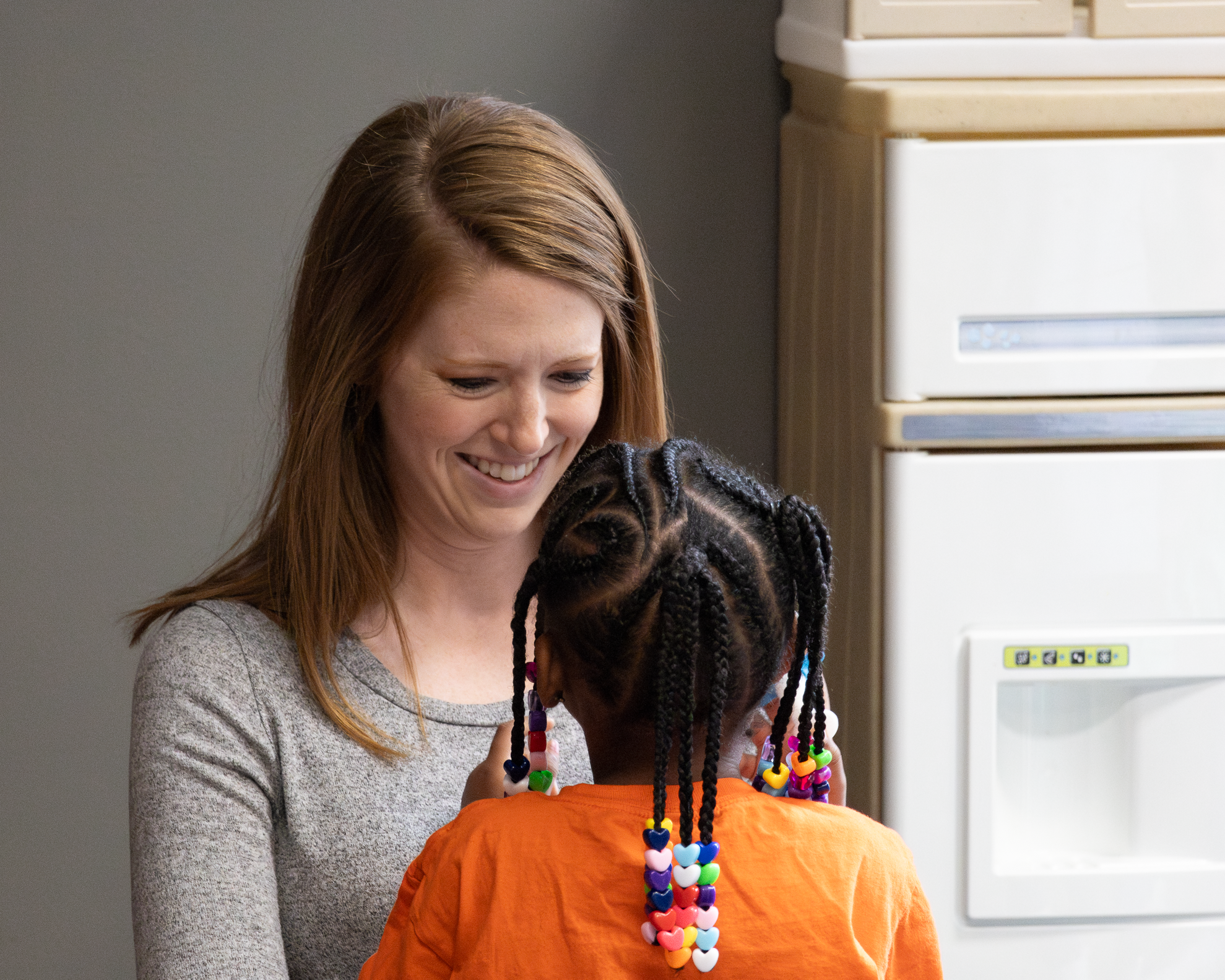 A woman with red hair smiling as she embraces a young girl with colorful beaded hair ties, against a plain wall and a white appliance in the background.