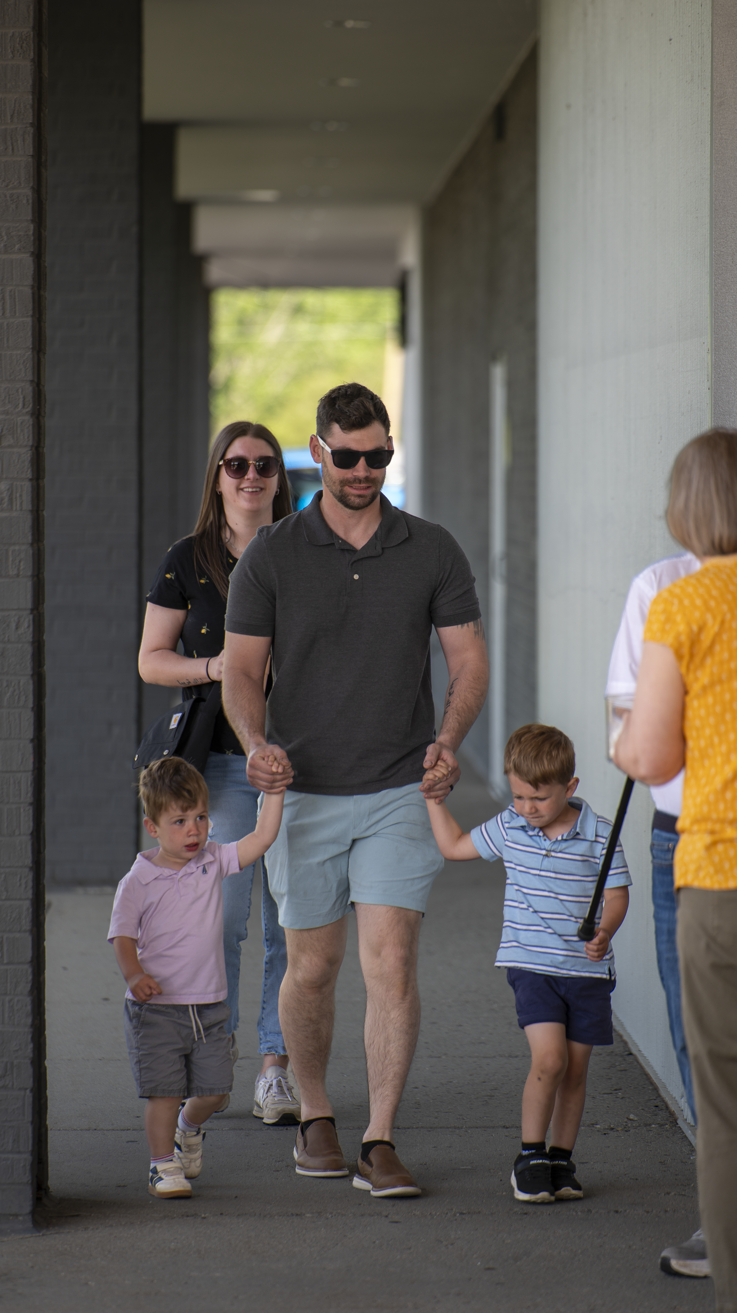 A man with sunglasses and a gray polo shirt walking with two young boys holding his hands, alongside a woman and other children in a shaded outdoor corridor.