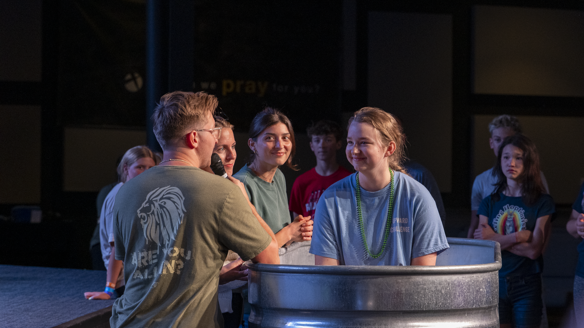 Group of young people, some smiling, gathered around a metal baptismal font on stage, during a church or youth event.