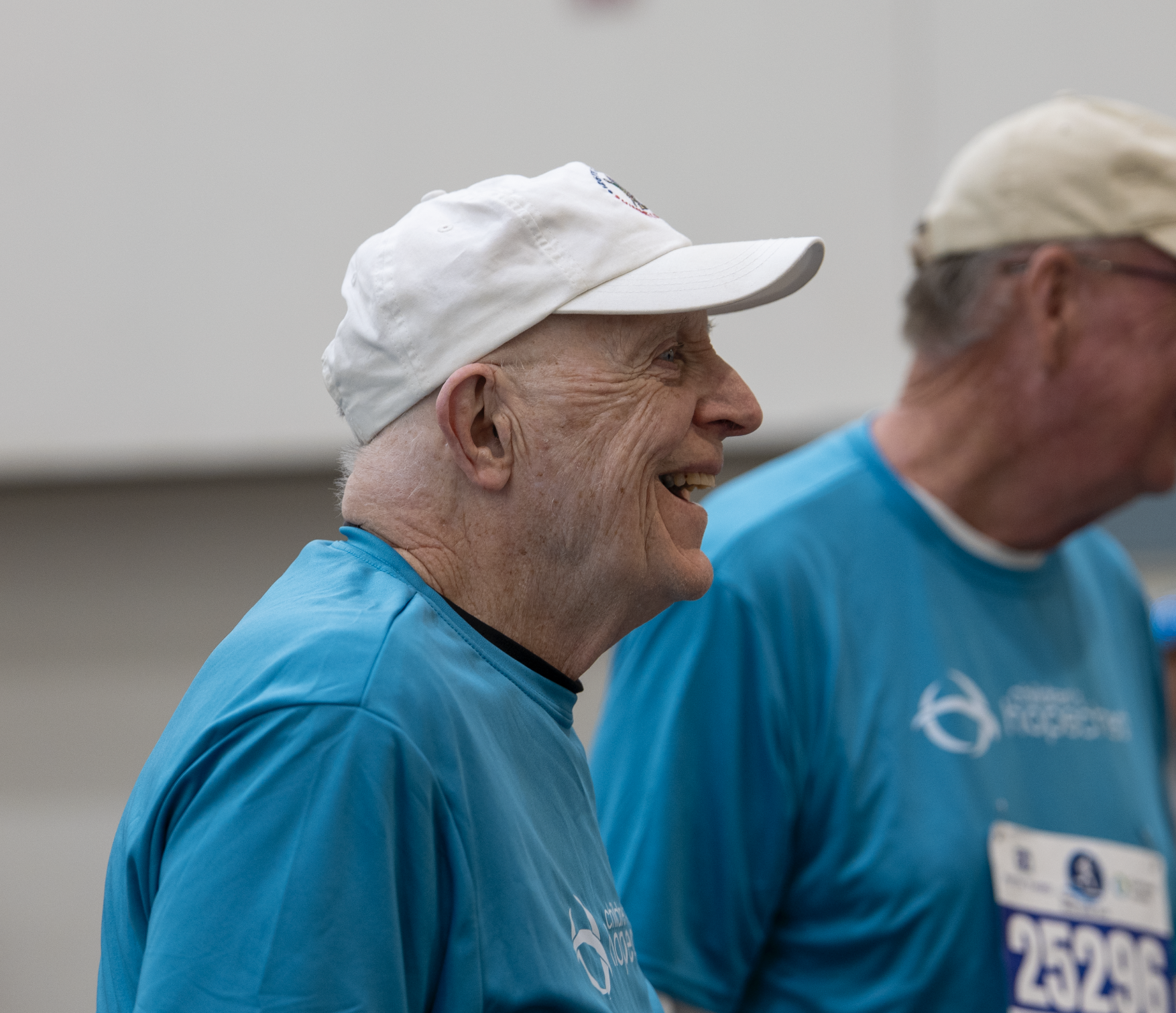 A smiling elderly man wearing a white baseball cap and a blue T-shirt, standing indoors alongside another person also in a blue T-shirt and white cap.