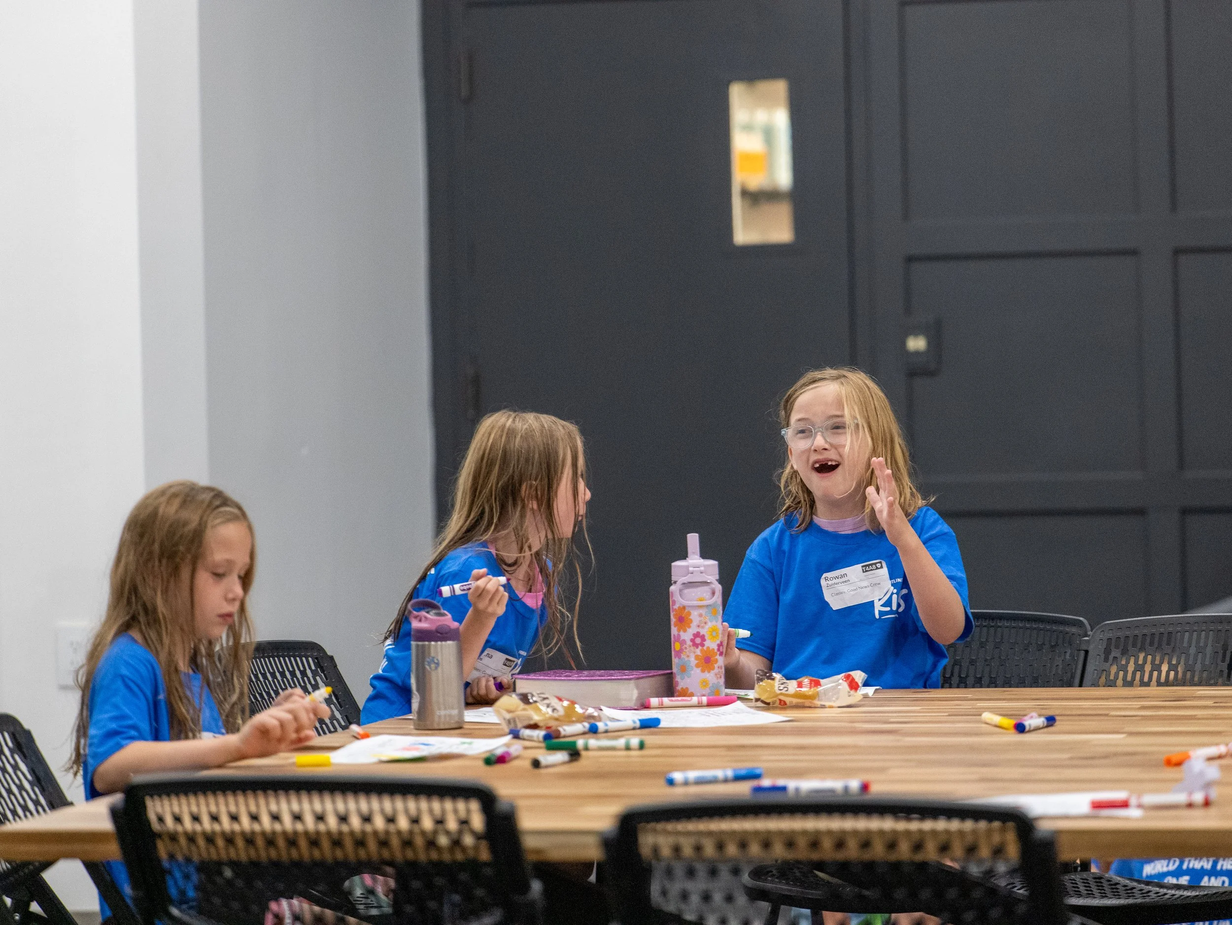 Four young girls with long hair sitting at a wooden table, engaging in arts and crafts with markers and paper, in a room with dark gray walls and a black door.